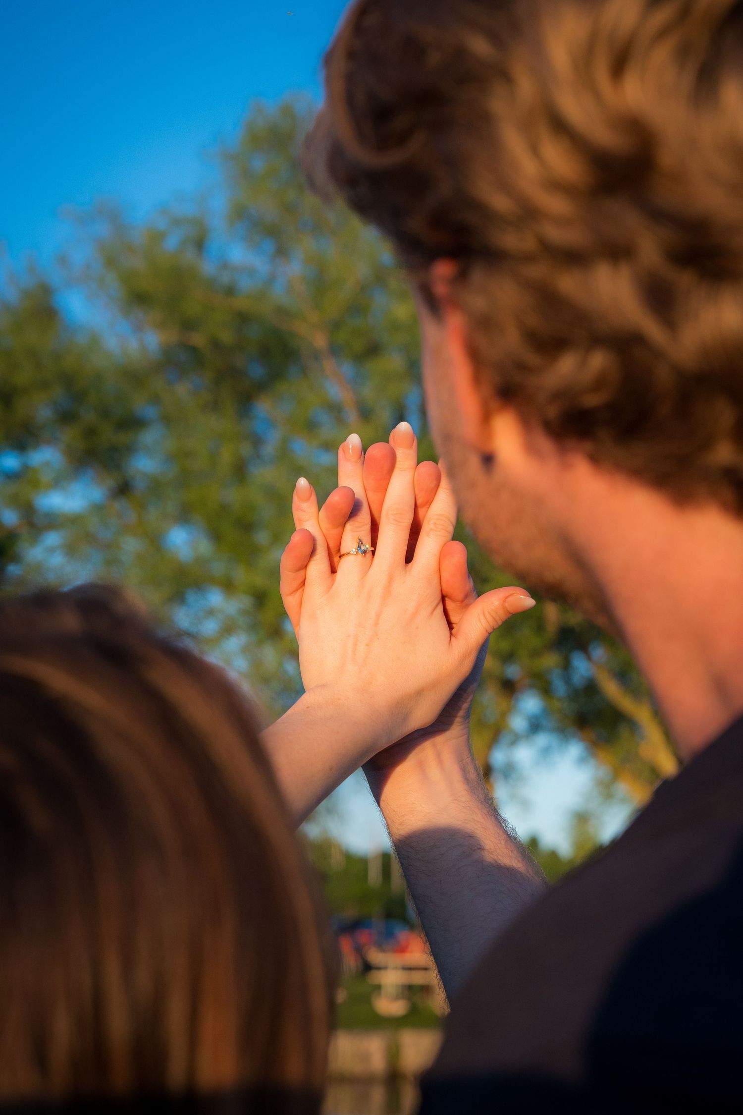 Close-up of an engagement ring on a woman's hand pressed against her partner's palm outdoors.