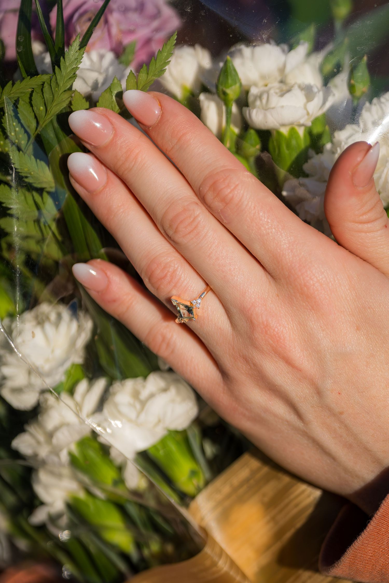 Close-up of a womans hand with elegant nude ombre nails wearing a delicate gold engagement ring, held against a bouquet.