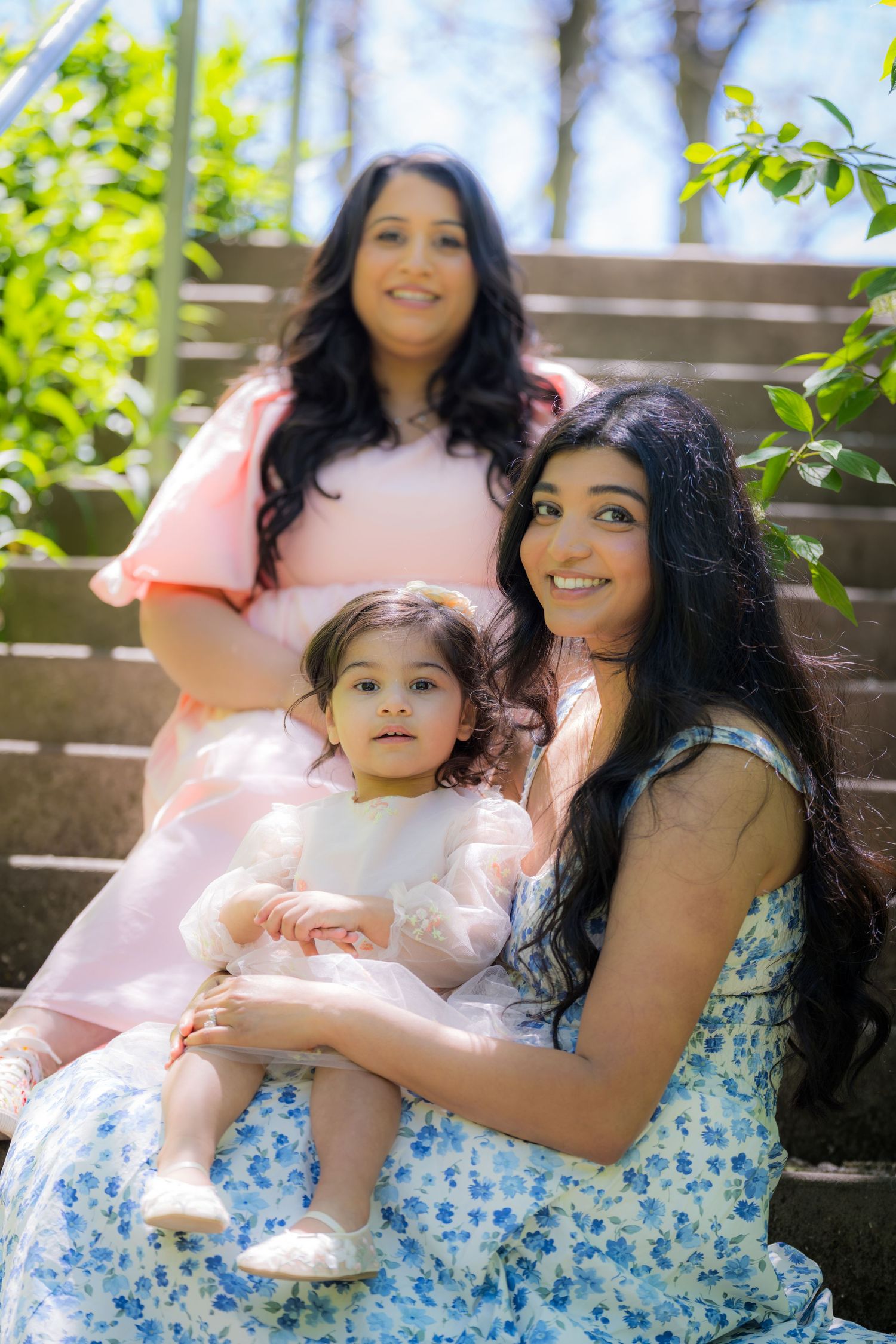 Two South Asian women and a toddler in white dress pose together on outdoor steps surrounded by greenery.