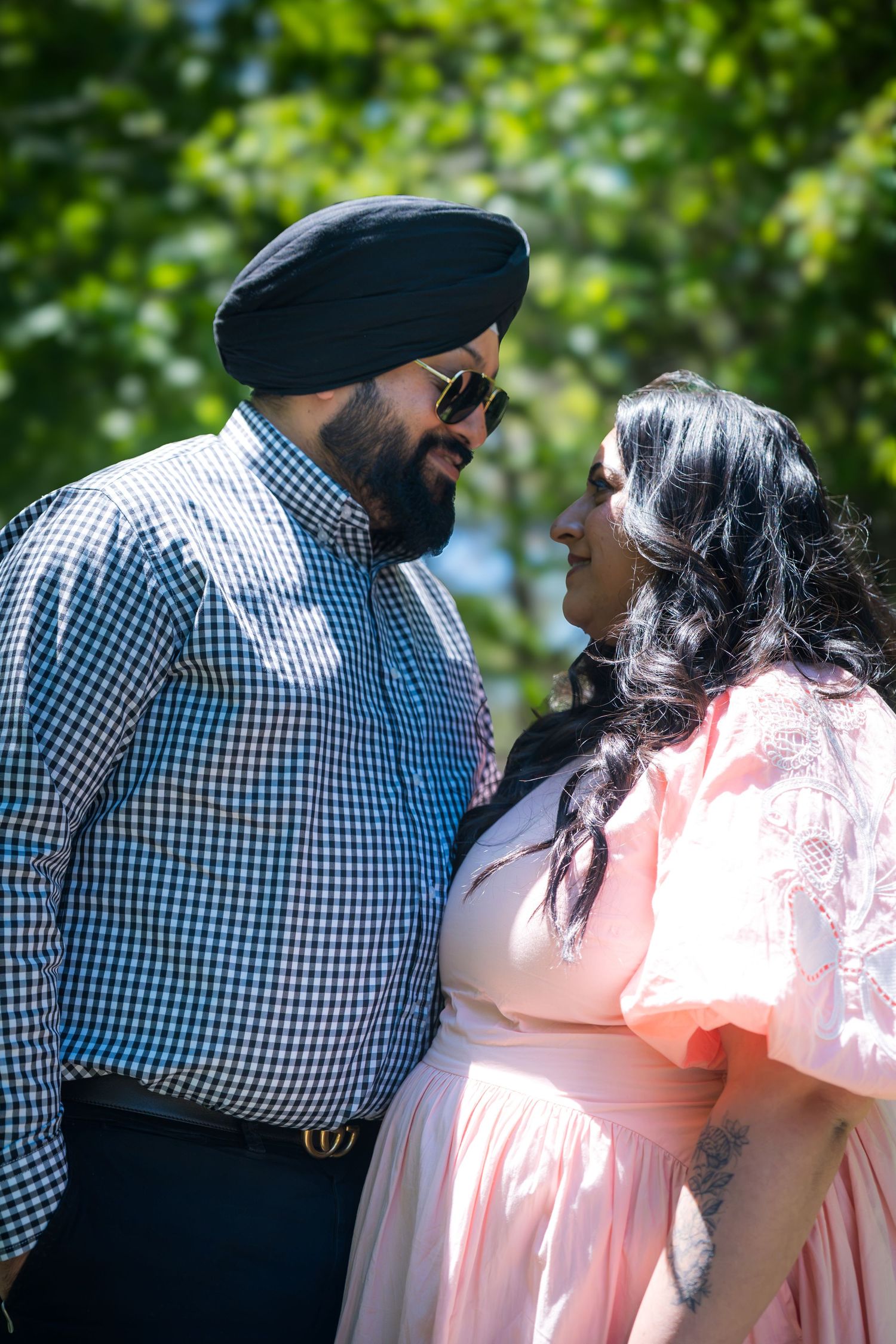 A Sikh man and woman in a pink dress share a romantic glance surrounded by lush green trees.