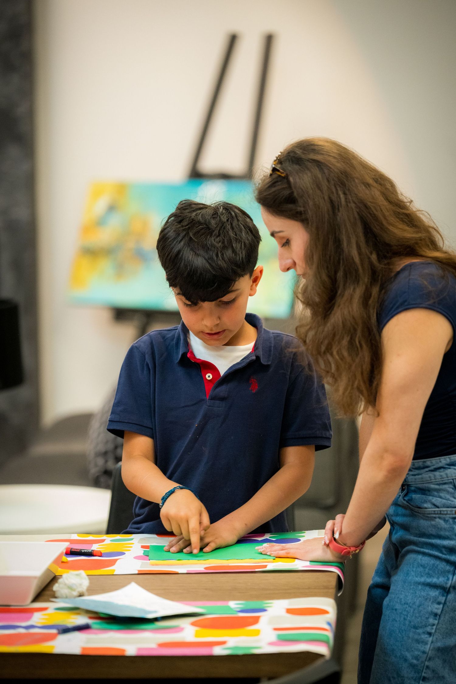 A woman guides a young boy in creating a vibrant paper collage artwork at a table.