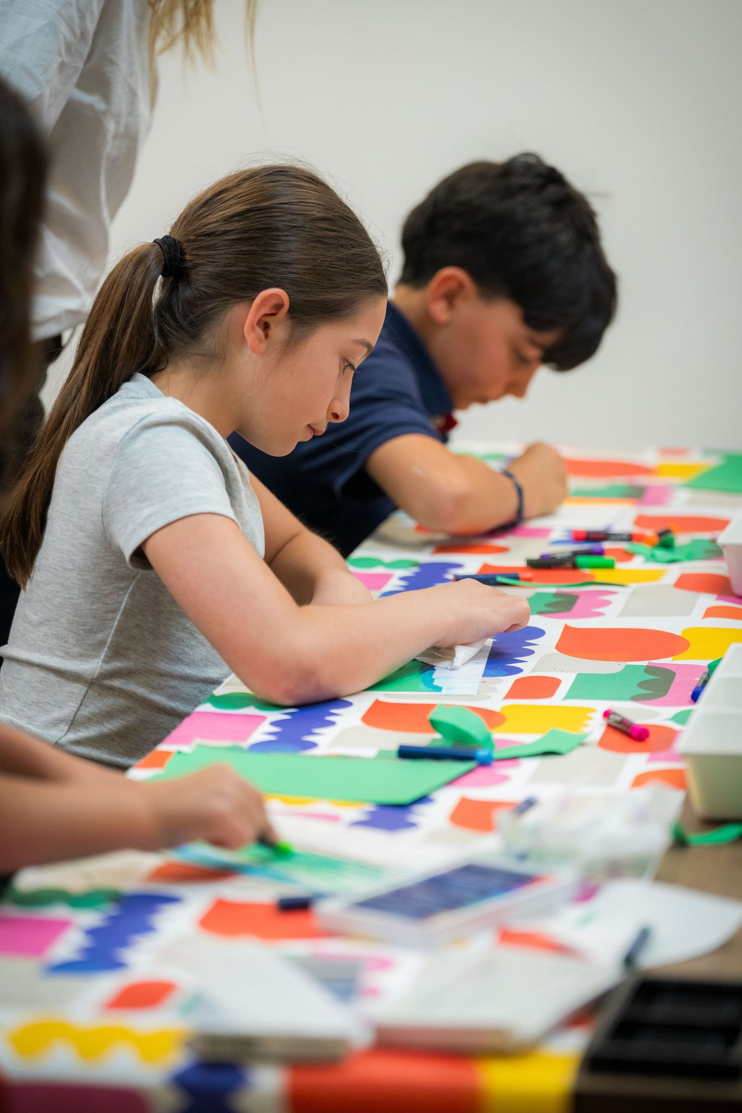 Girl with ponytail focuses on colorful paper craft project at vibrant art table during children's workshop.