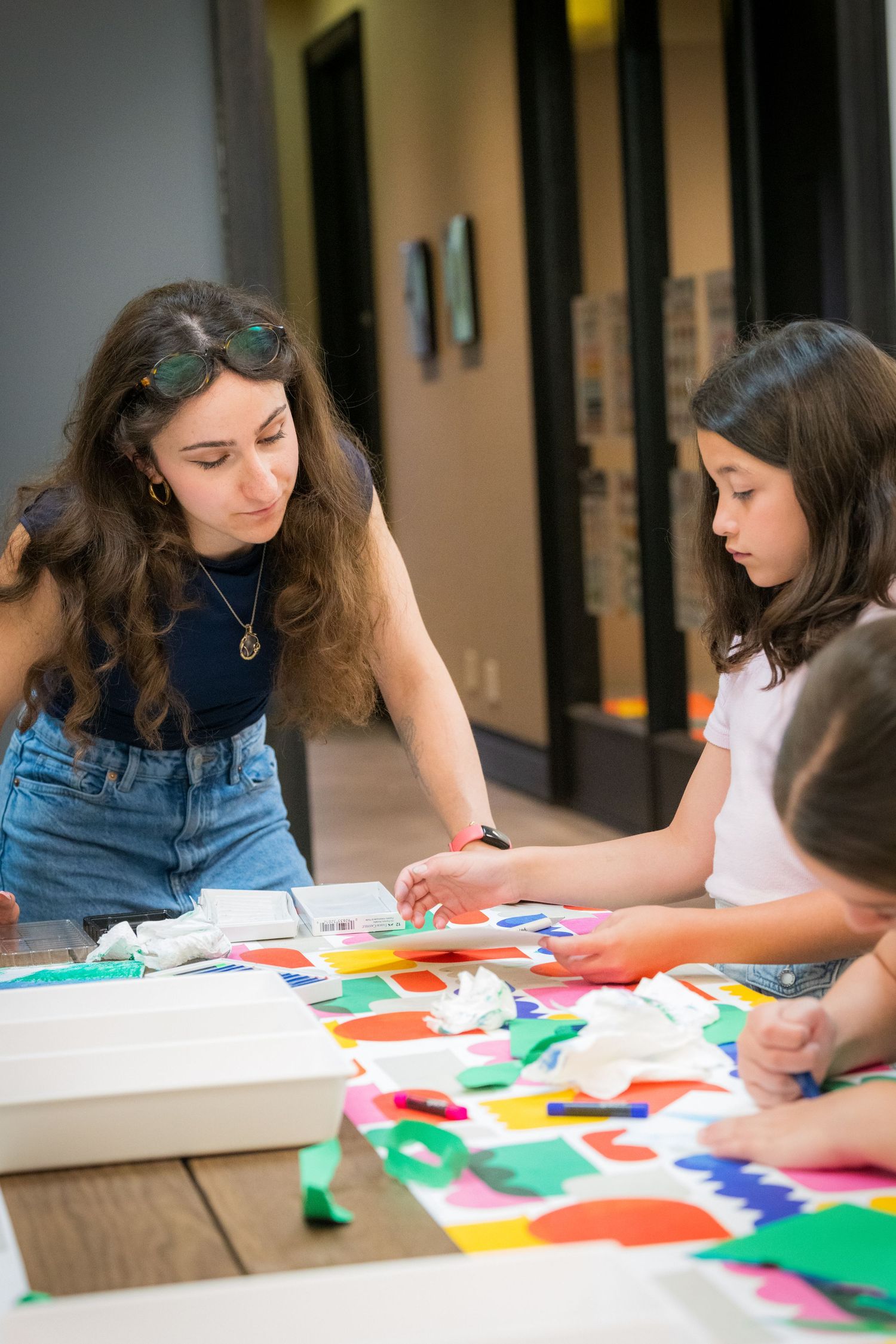 Female art instructor guides two girls working with colorful paper cutouts on large table.