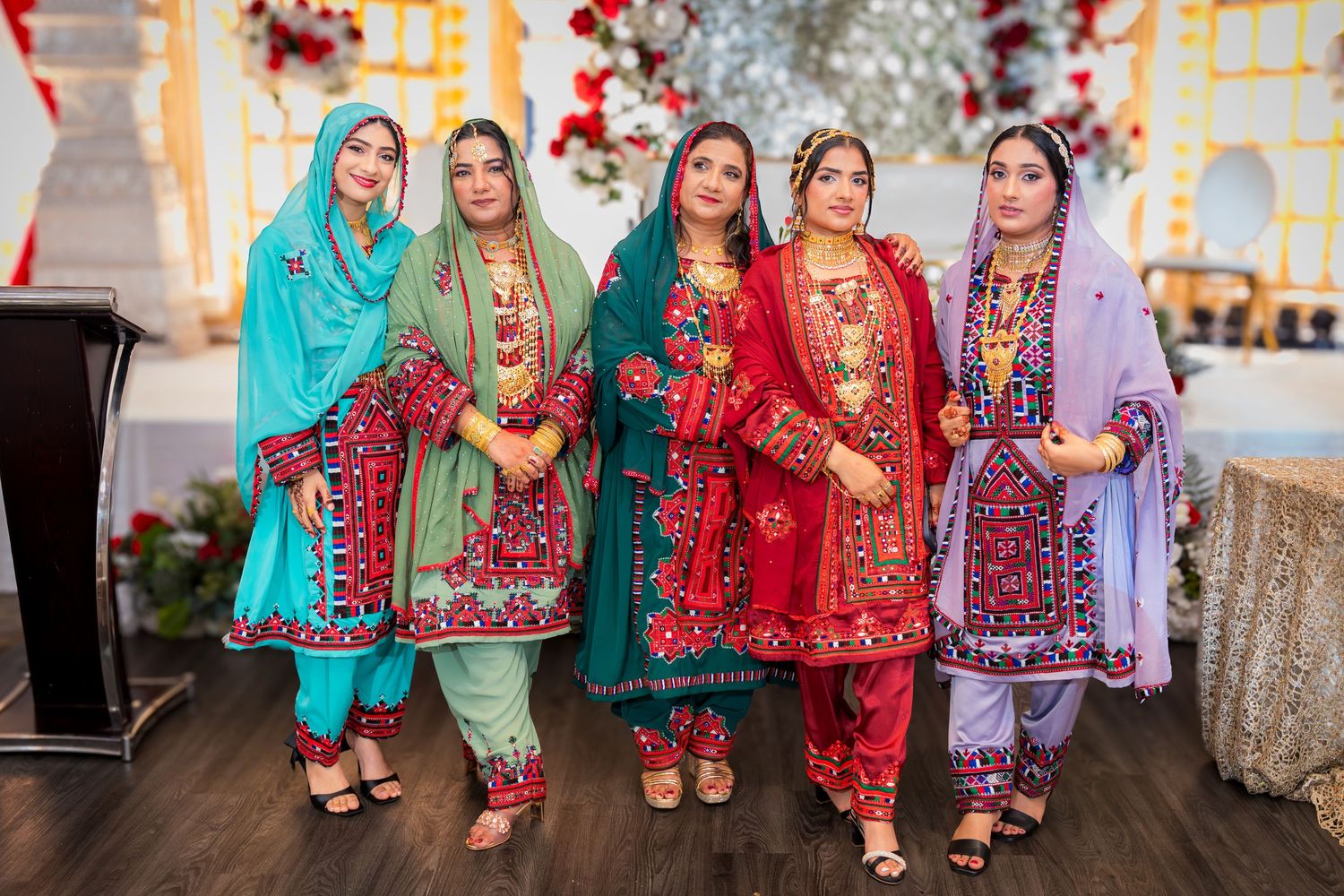 Five women in colorful traditional Pakistani outfits with matching dupattas pose together at a celebration.
