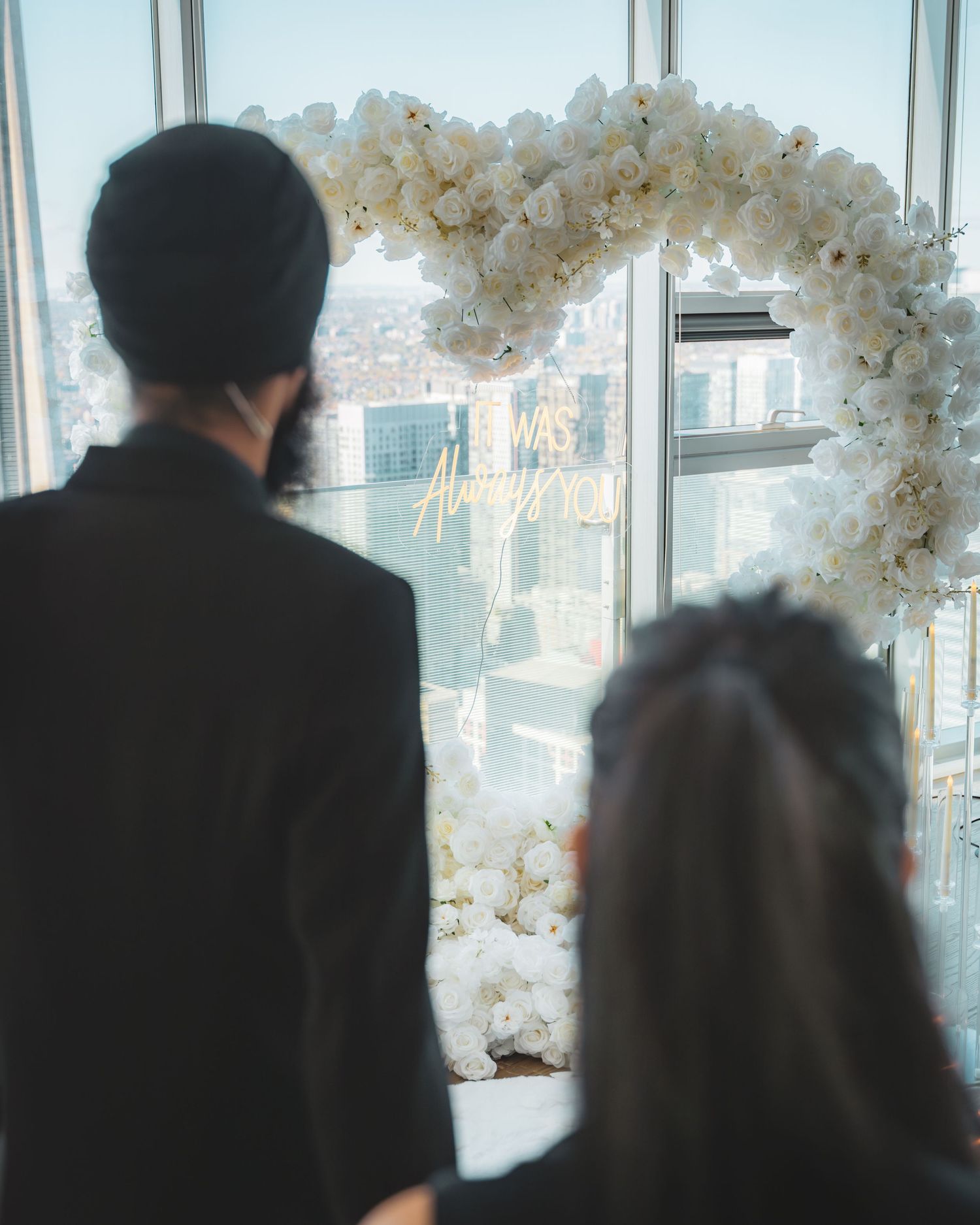 People in formal attire stand silhouetted against large windows with white floral decorations in an elegant venue.