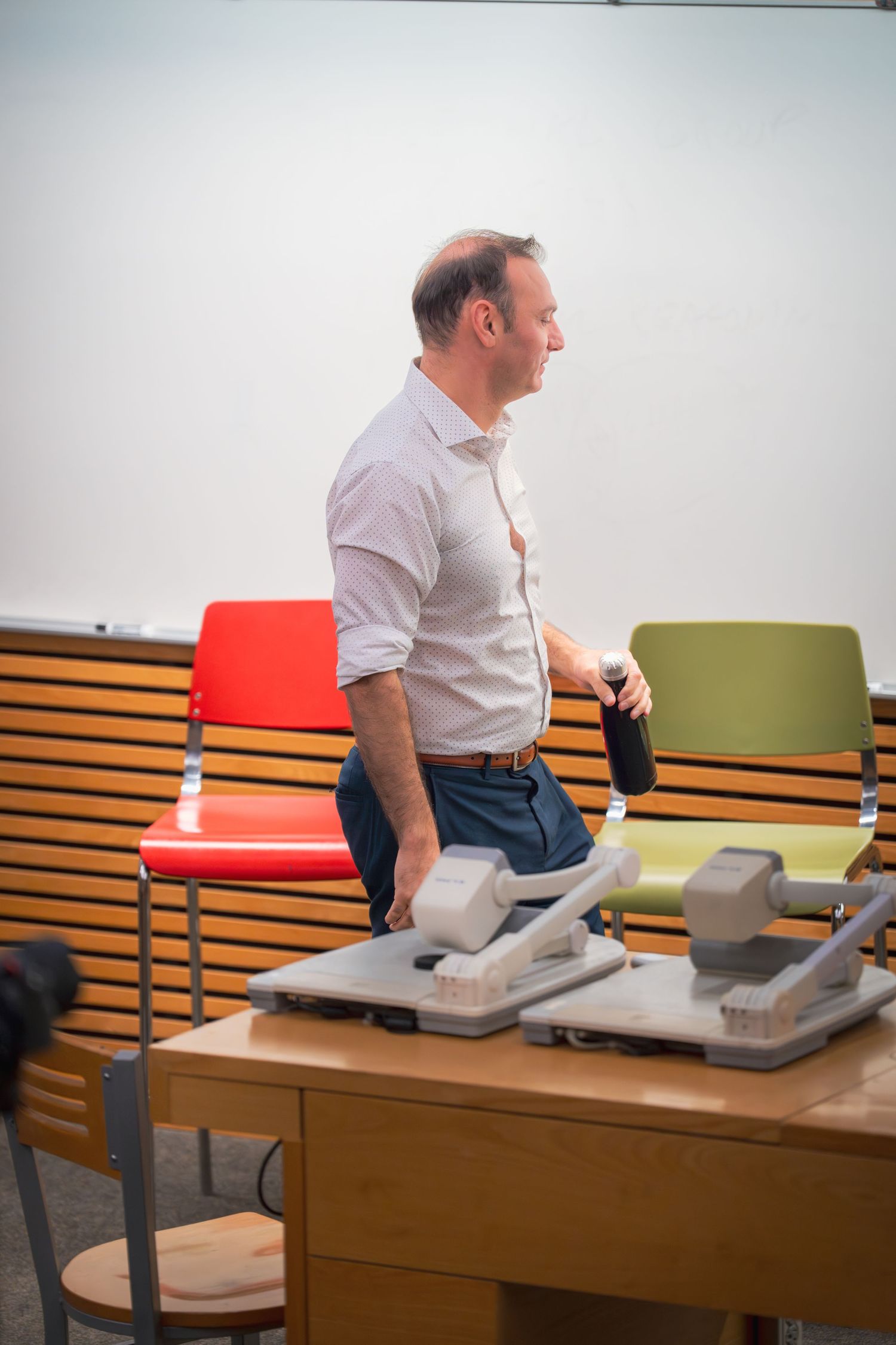 Speaker in white shirt gestures while seated at presentation desk with equipment.