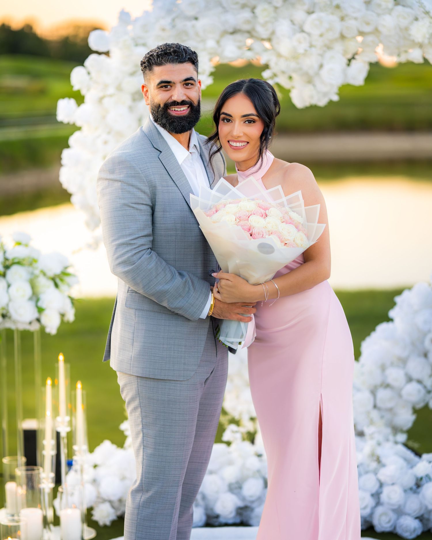Happy couple celebrating their engagement under white floral arch with sunset backdrop.