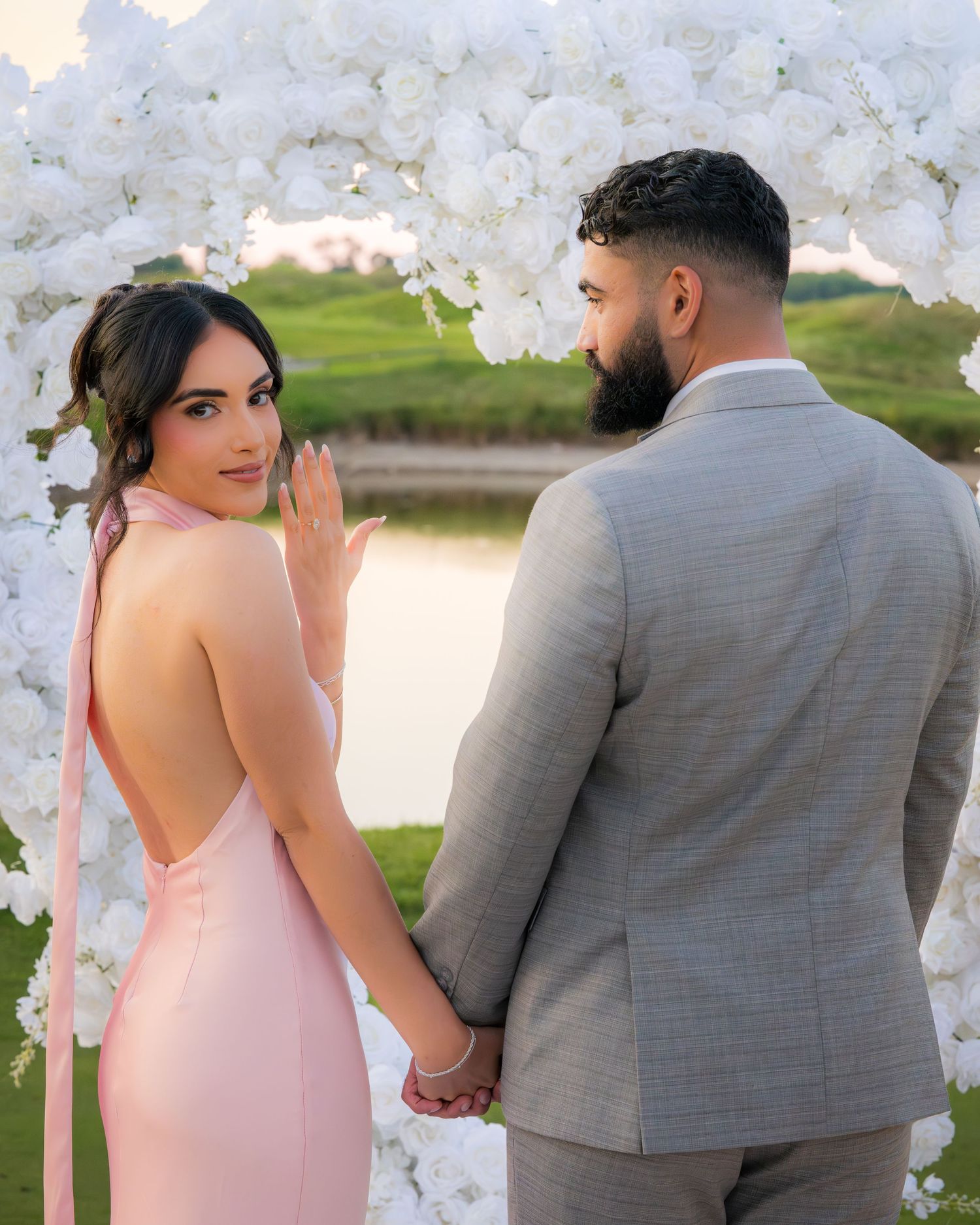 Elegant couple stands together near white floral arrangements on golf course.