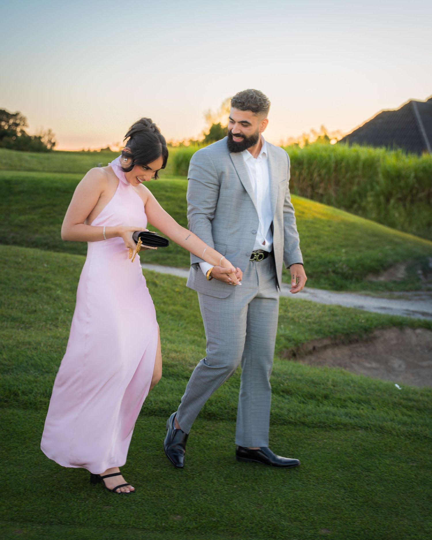 Couple in formal attire walking along grassy path during sunset photography session.