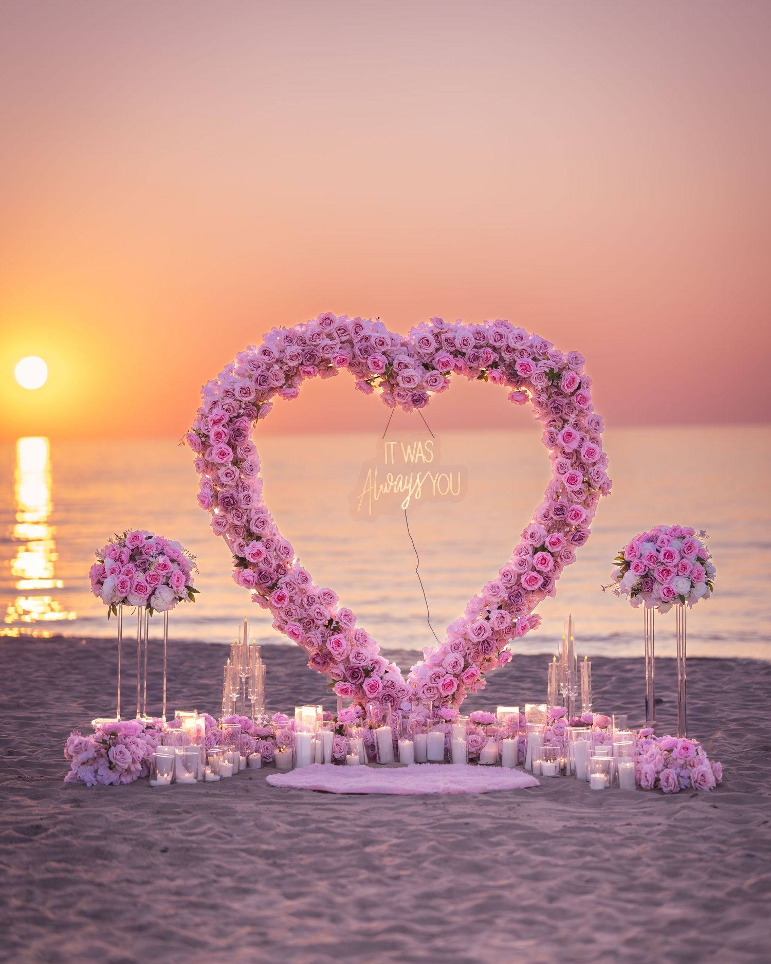 Heart-shaped pink floral arch on beach at sunset with glowing orange sky and ocean backdrop.