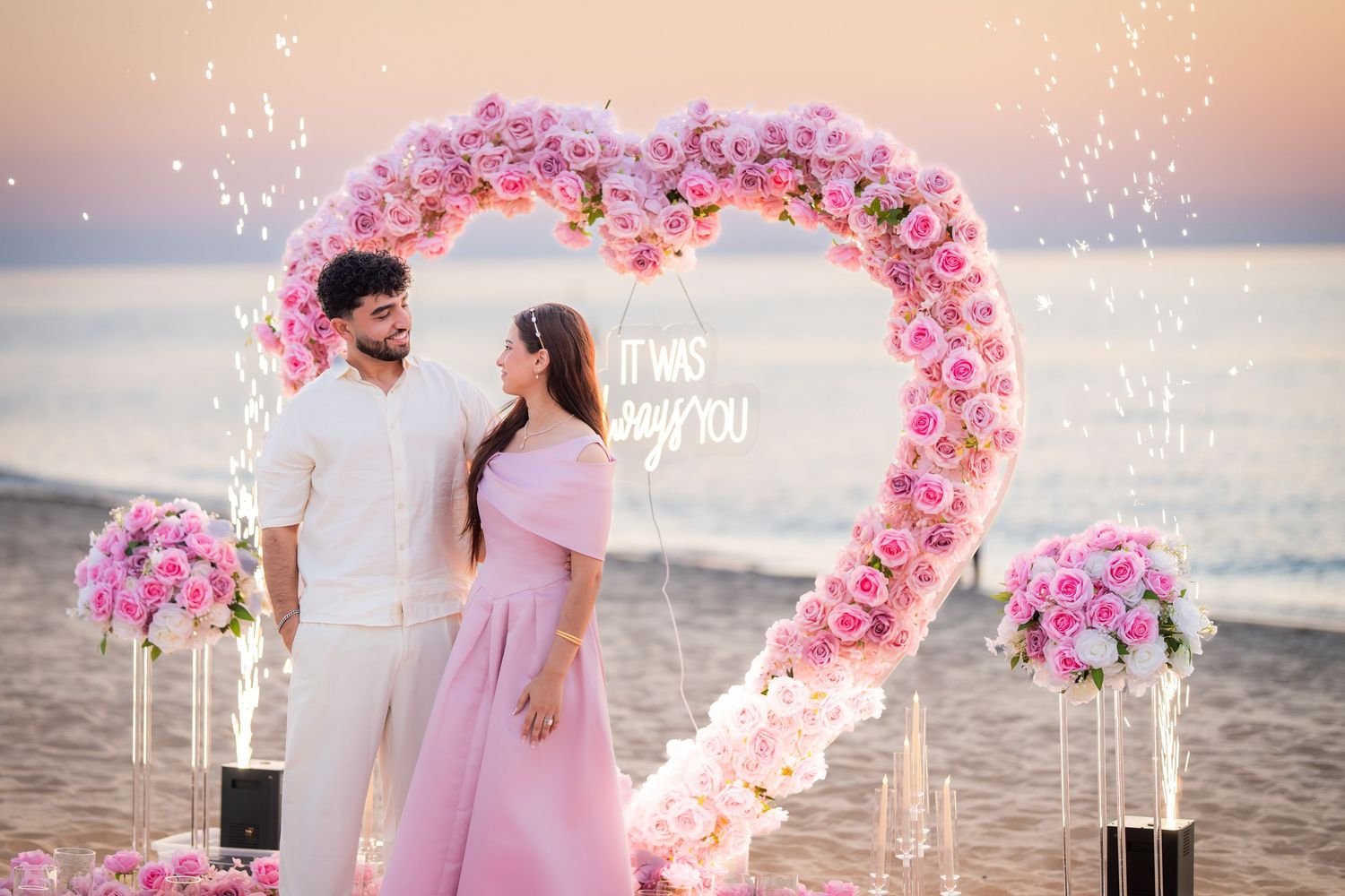 Pink floral heart arch on beach with sparklers creating magical atmosphere.