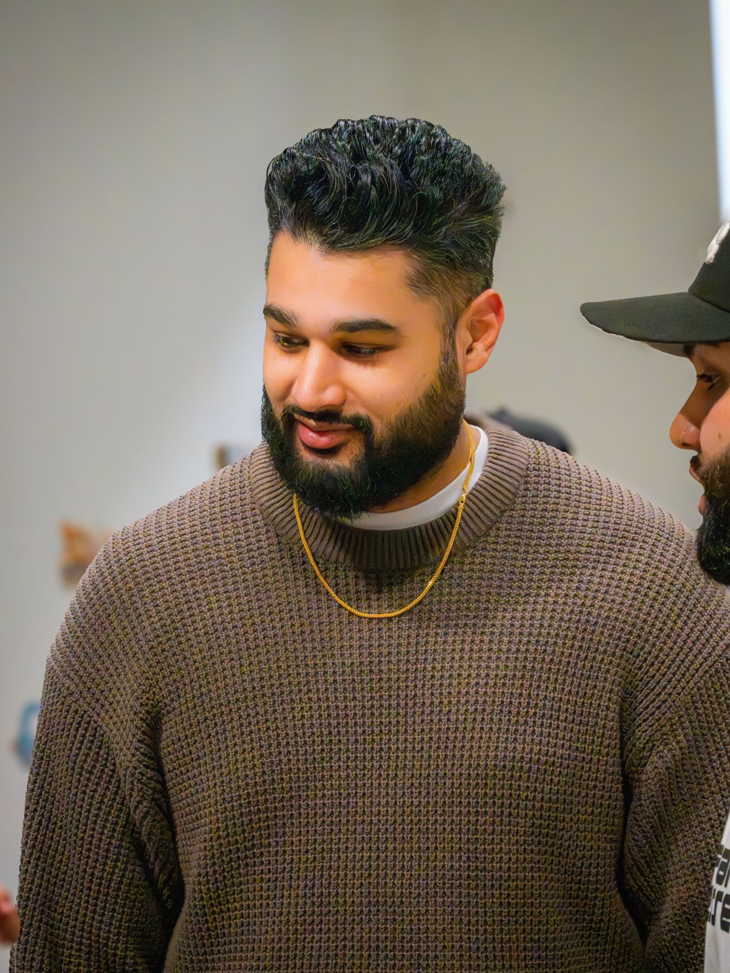 Bearded man in brown waffle knit sweater with gold chain smiling at indoor gathering.