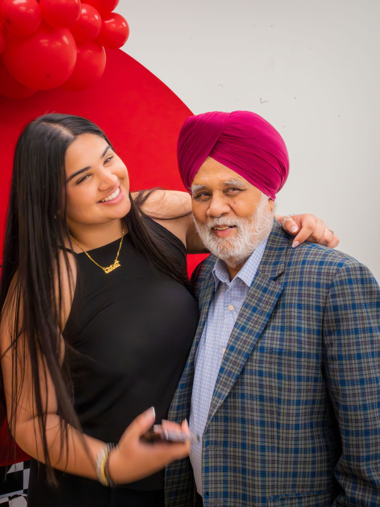 A young woman and an elderly Sikh man wearing a magenta turban smile together in front of red balloons.