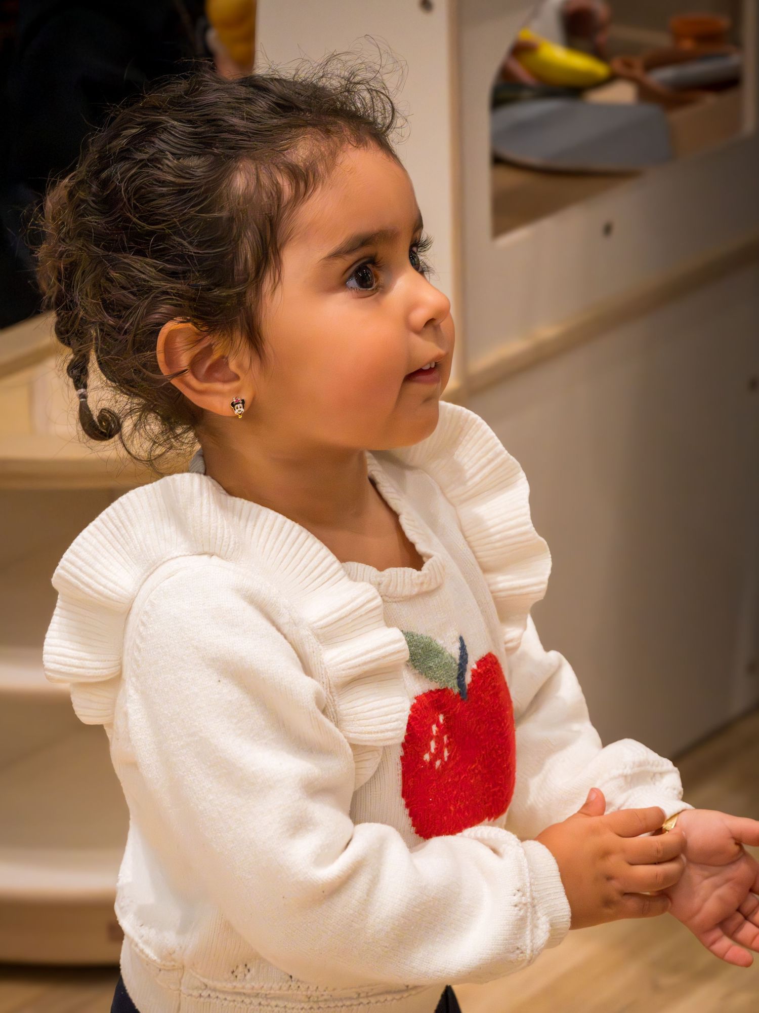 Little girl in white ruffled apple sweater standing near a wooden play kitchen.