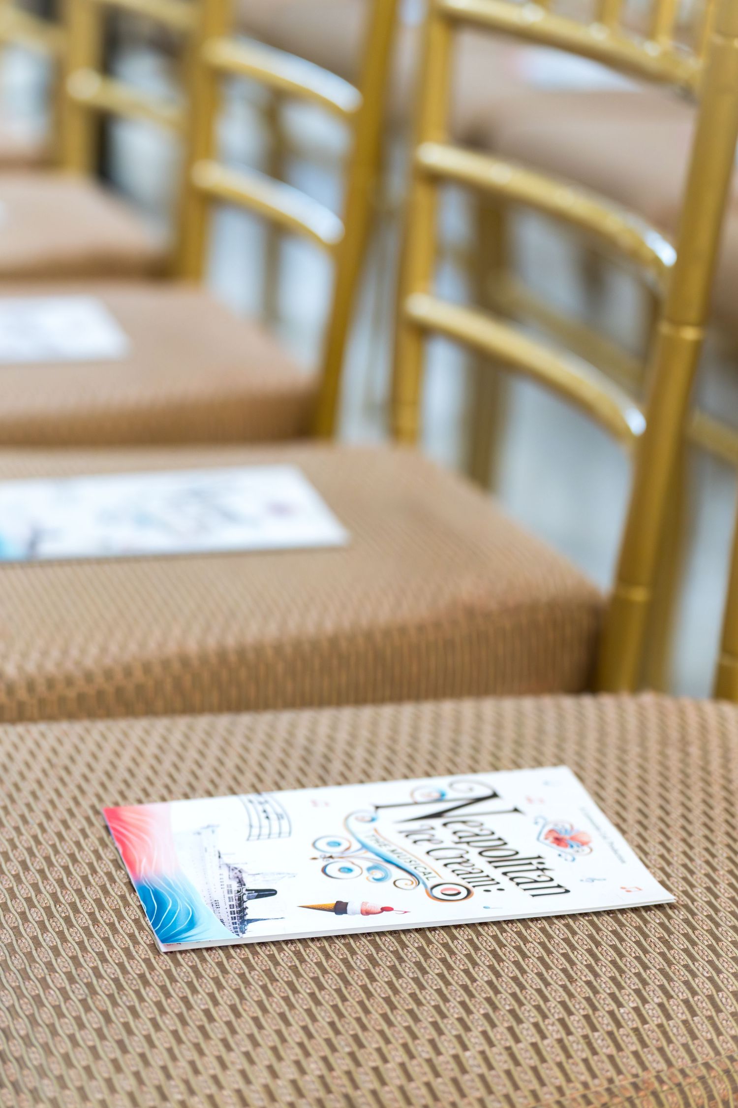 Gold chiavari chairs with colorful wedding ceremony programs placed on tan cushioned seats at an elegant event venue.