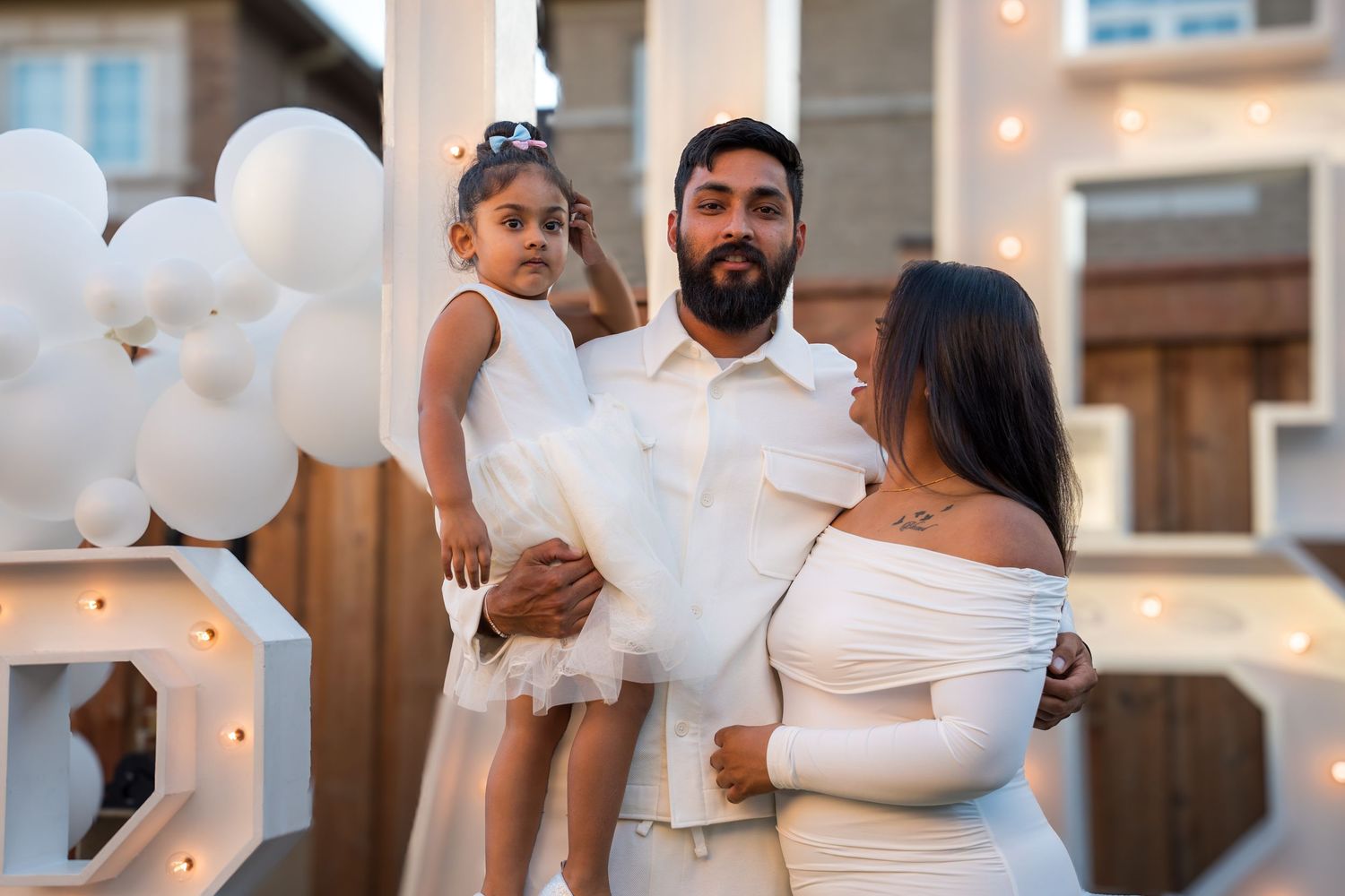 A family of three in white outfits poses together at an elegant outdoor baby shower event.