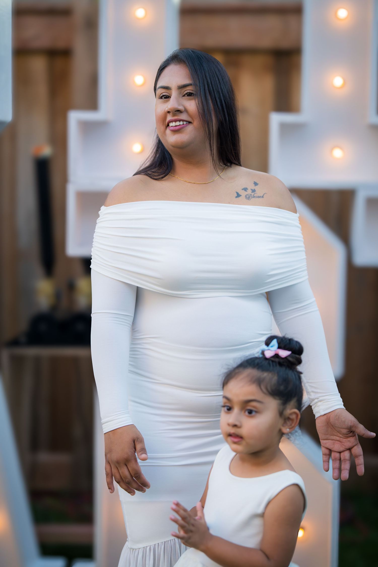 Pregnant woman in white off-shoulder dress posing with young girl at a baby shower celebration.