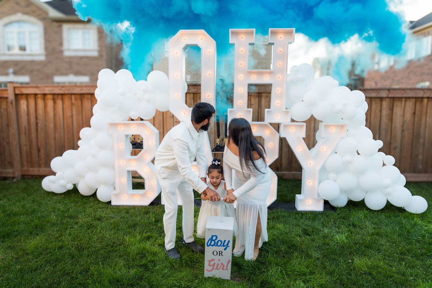 Couple with toddler reveal baby gender with blue smoke at outdoor OH BABY marquee letter party setup.