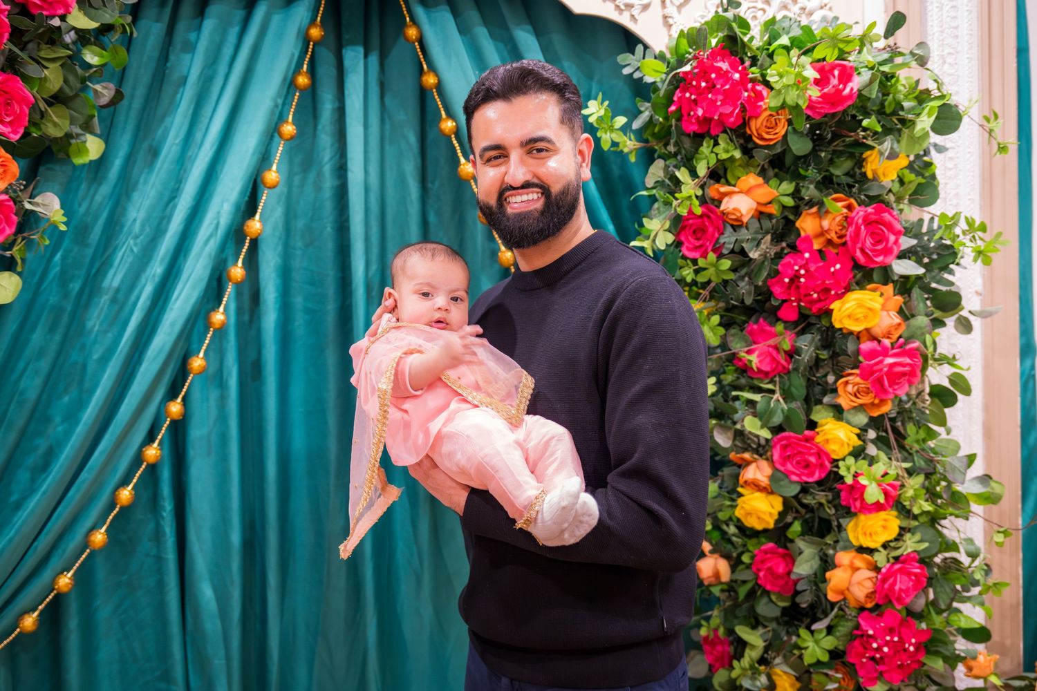 Bearded man in black outfit proudly holding pink-dressed newborn before colorful floral display.
