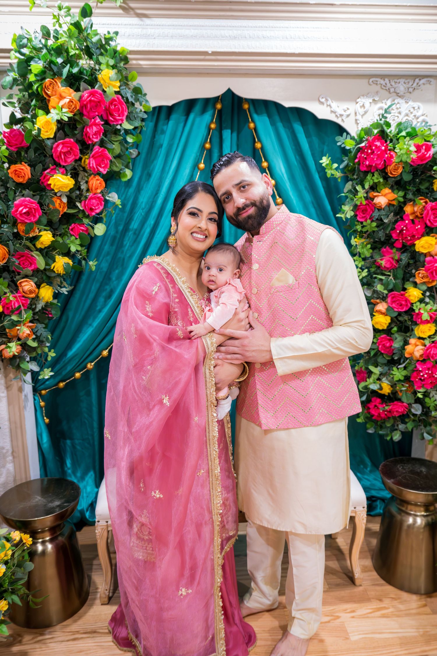 Indian couple in pink traditional attire holding baby at decorated ceremony with colorful floral backdrop.