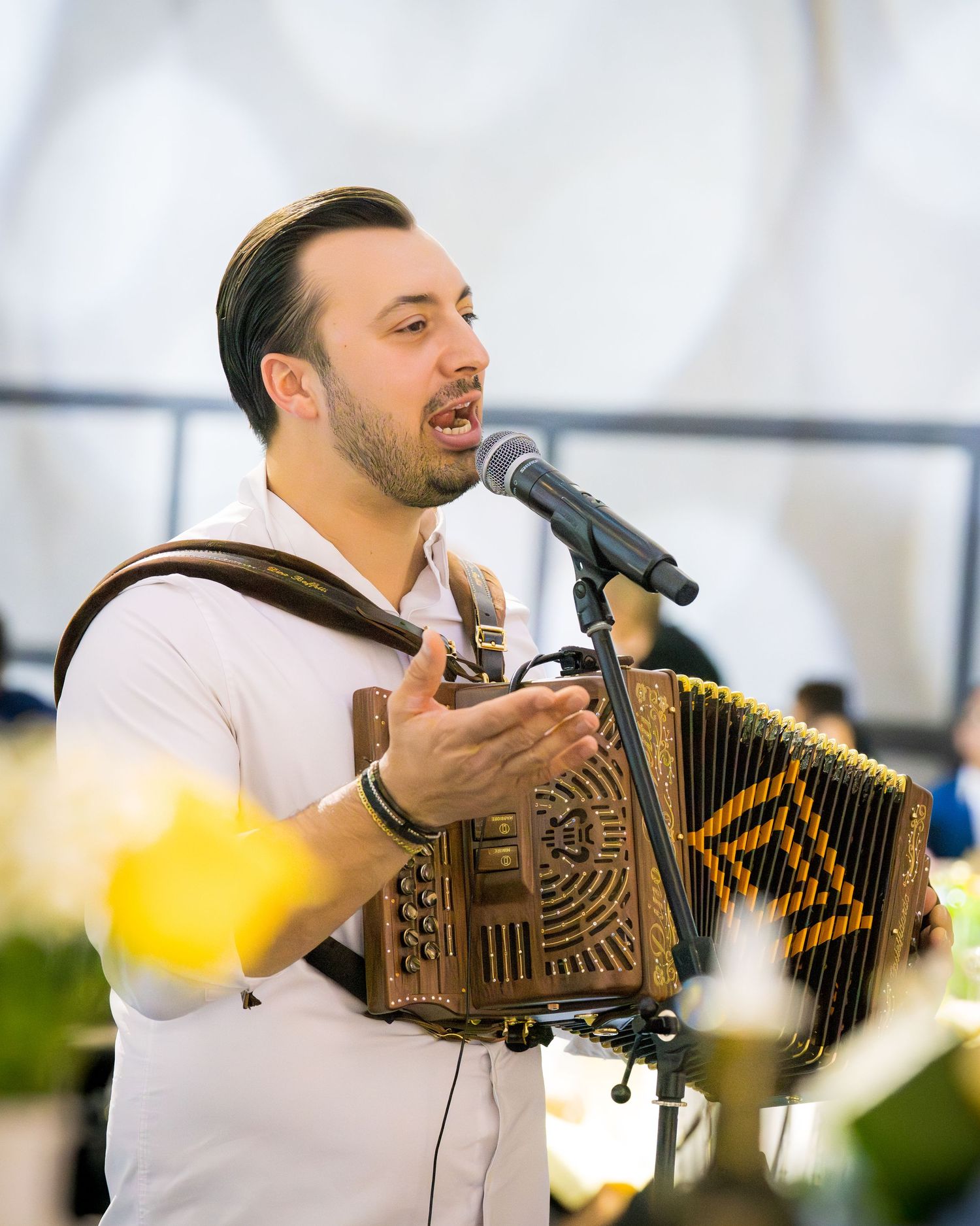 Male musician singing and playing ornate accordion at microphone during indoor celebration event.