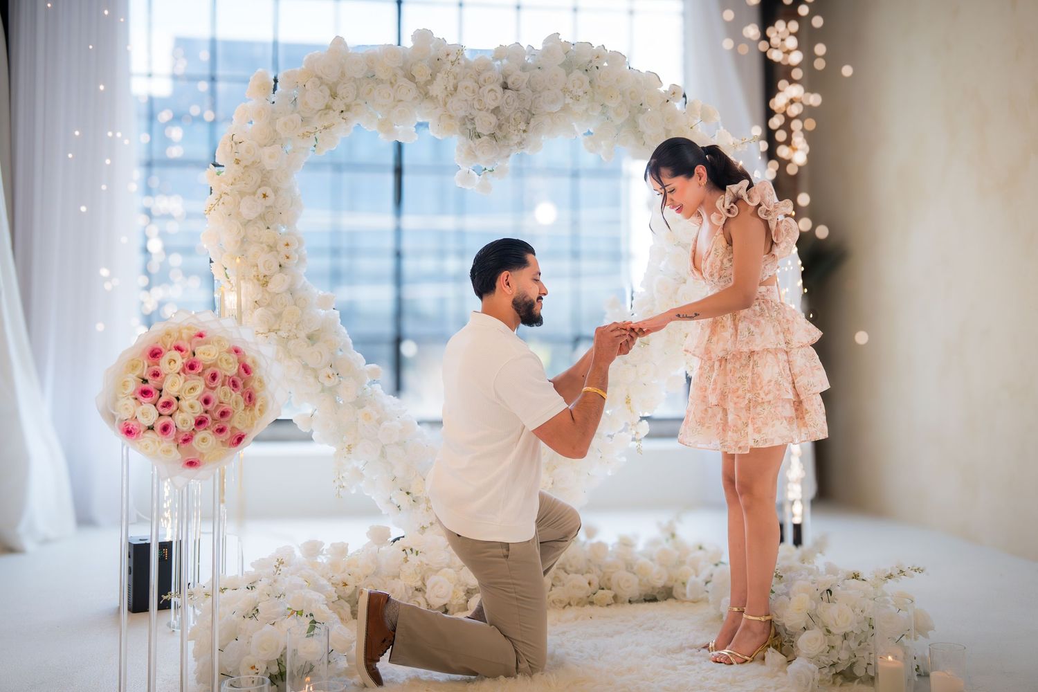 Man proposes to woman on one knee in front of a large white floral heart arch with romantic decor.