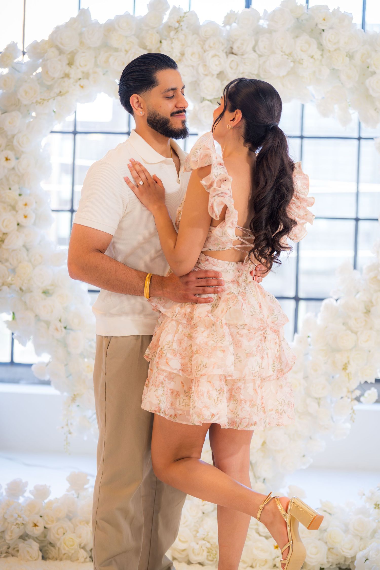 Couple embracing and gazing at each other before heart-shaped white floral arch during proposal event.
