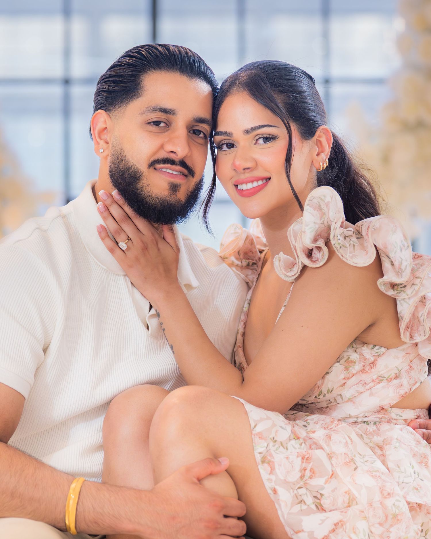 Newly engaged couple posing closely together smiling with white floral decorations in background.