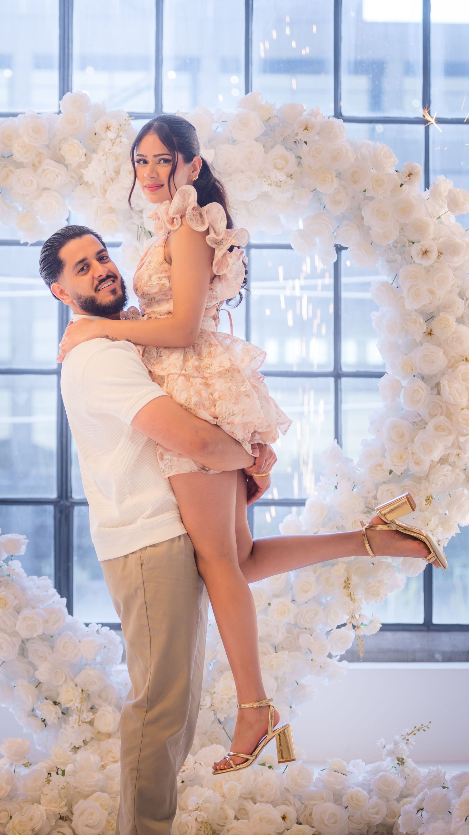 Couple poses together in front of white floral heart arch with sparklers.