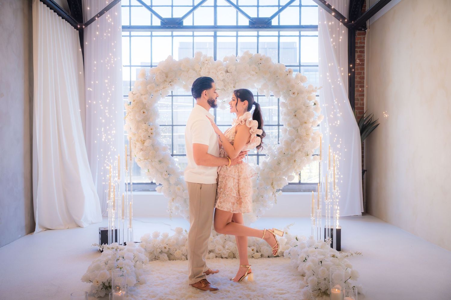 Couple dancing together beneath a stunning white floral heart arch with glowing lights.