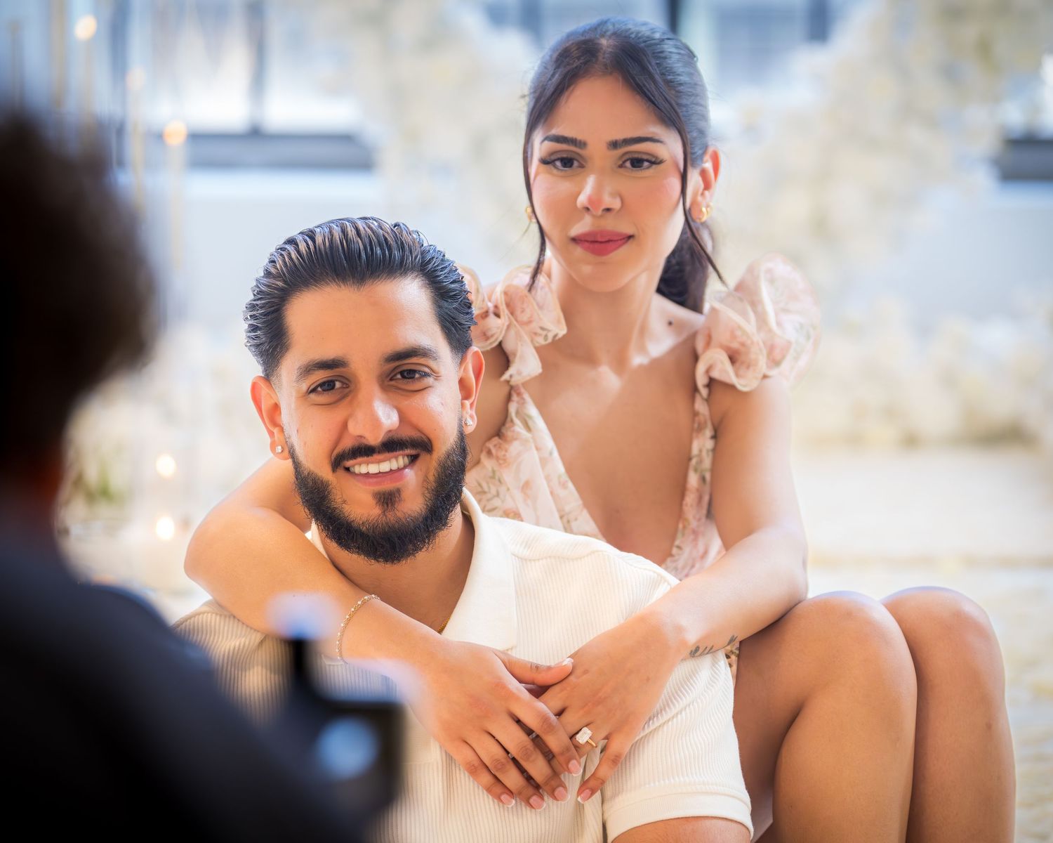 Newly engaged couple posing for photo, woman leaning on mans shoulder showing engagement ring.