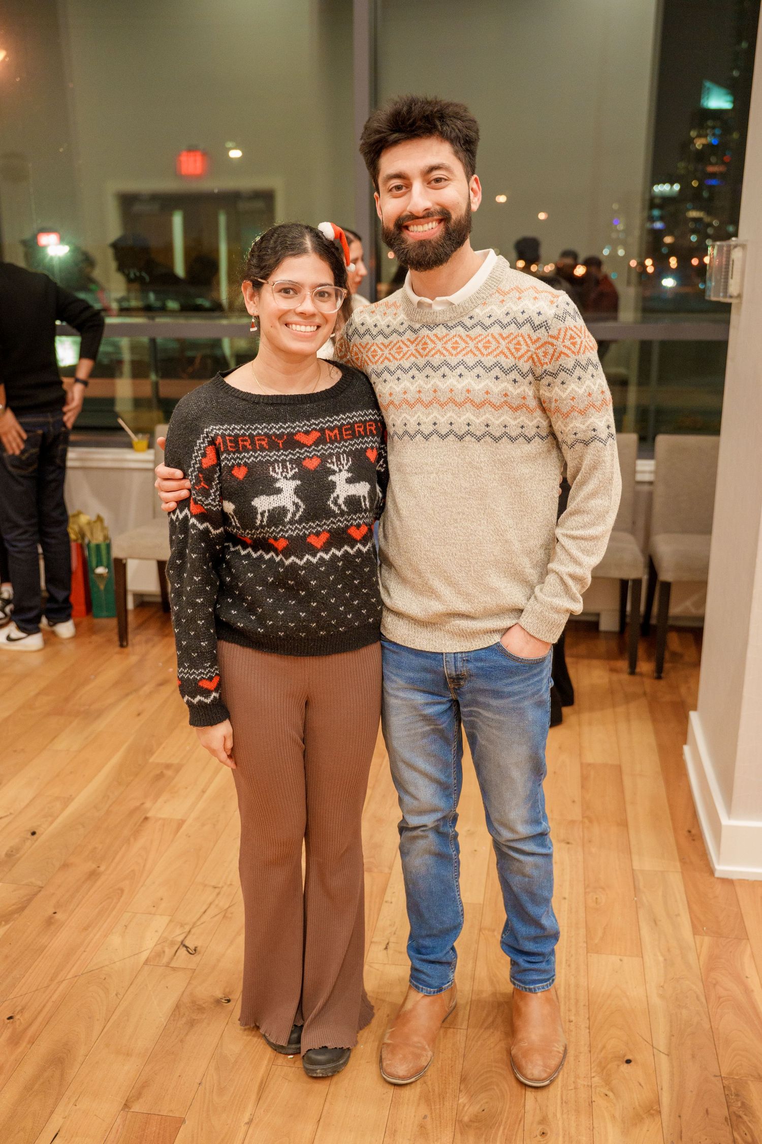 People wearing festive holiday sweaters pose together at an indoor gathering with hardwood floors.