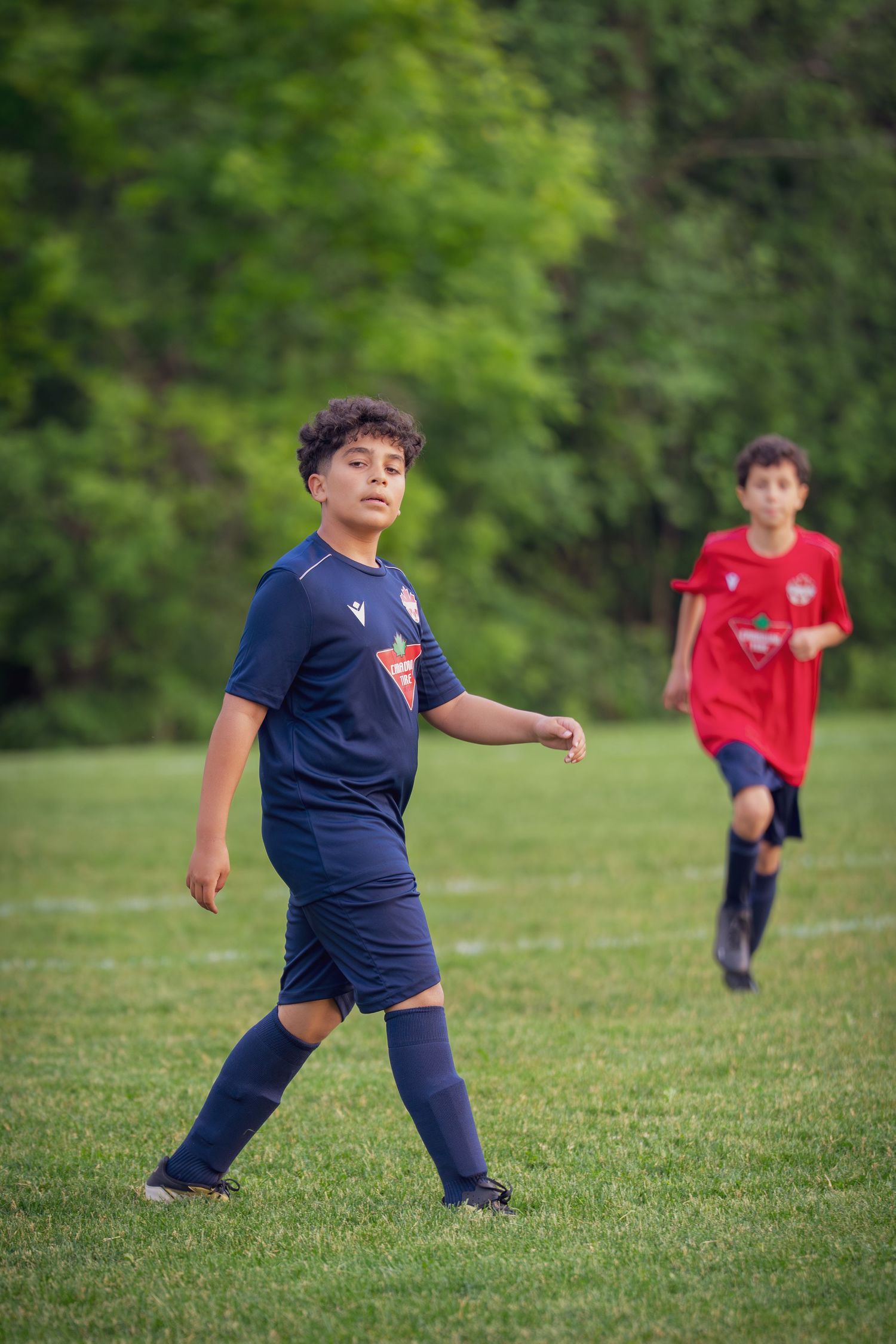 Two children in sports uniforms practice soccer moves on a grassy field with trees in the background.