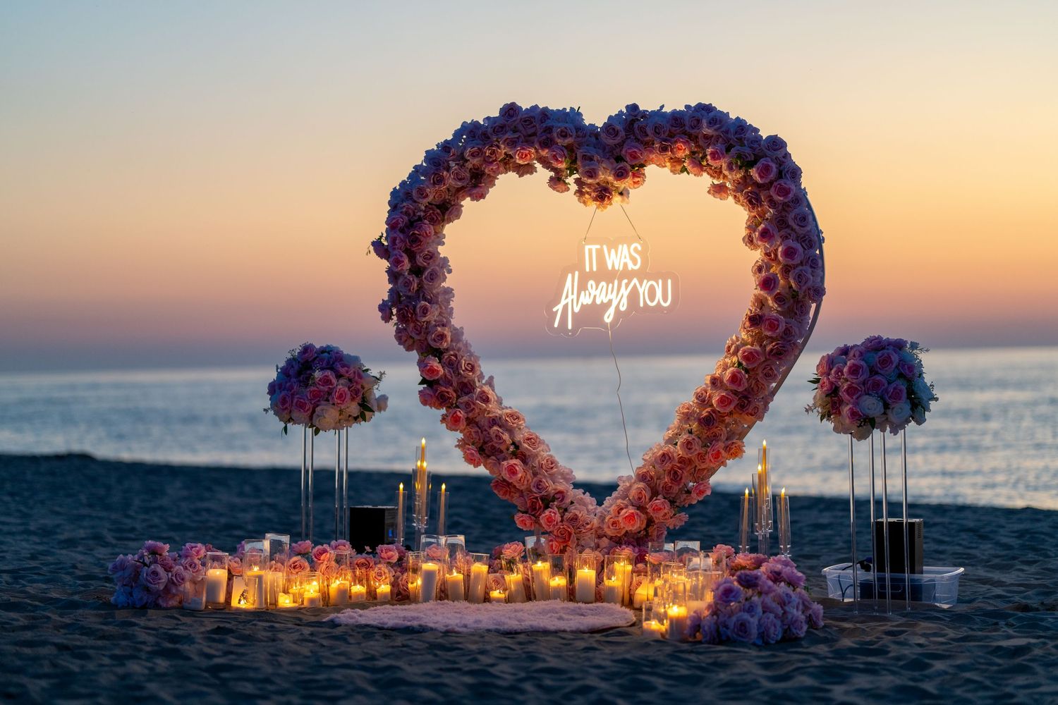 Pink floral heart arch glowing against sunset sky over ocean waters.
