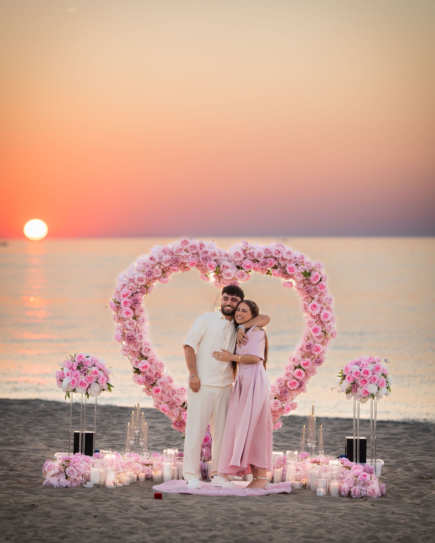 Beachside marriage proposal scene with pink flower arch and lanterns at dusk.
