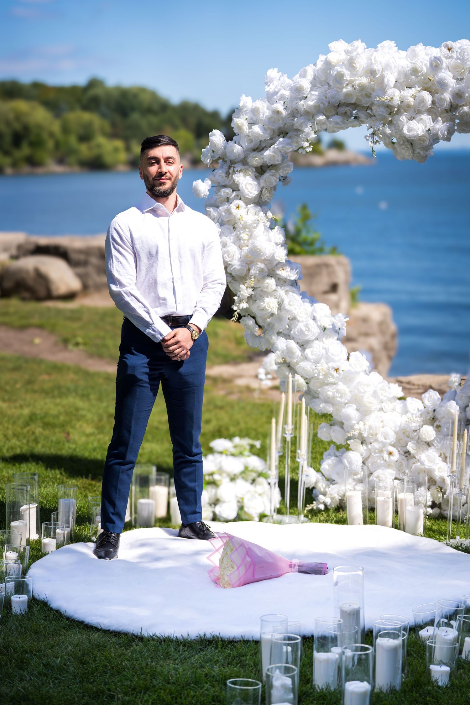 An elegant waterfront wedding setup with white balloon arches and glass candle holders on the grass.