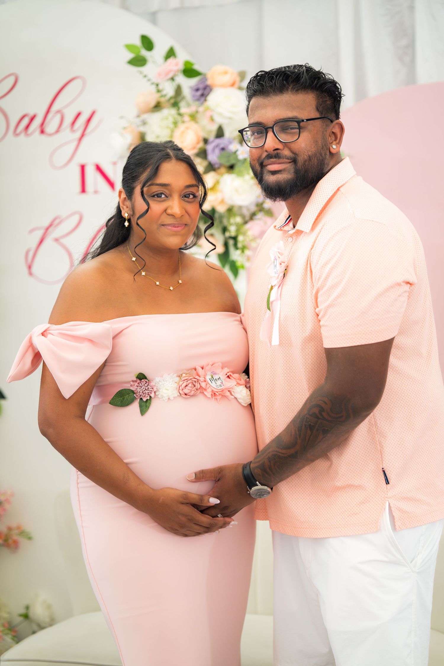 A couple shares tender moments in front of a floral wall at what appears to be a baby shower.