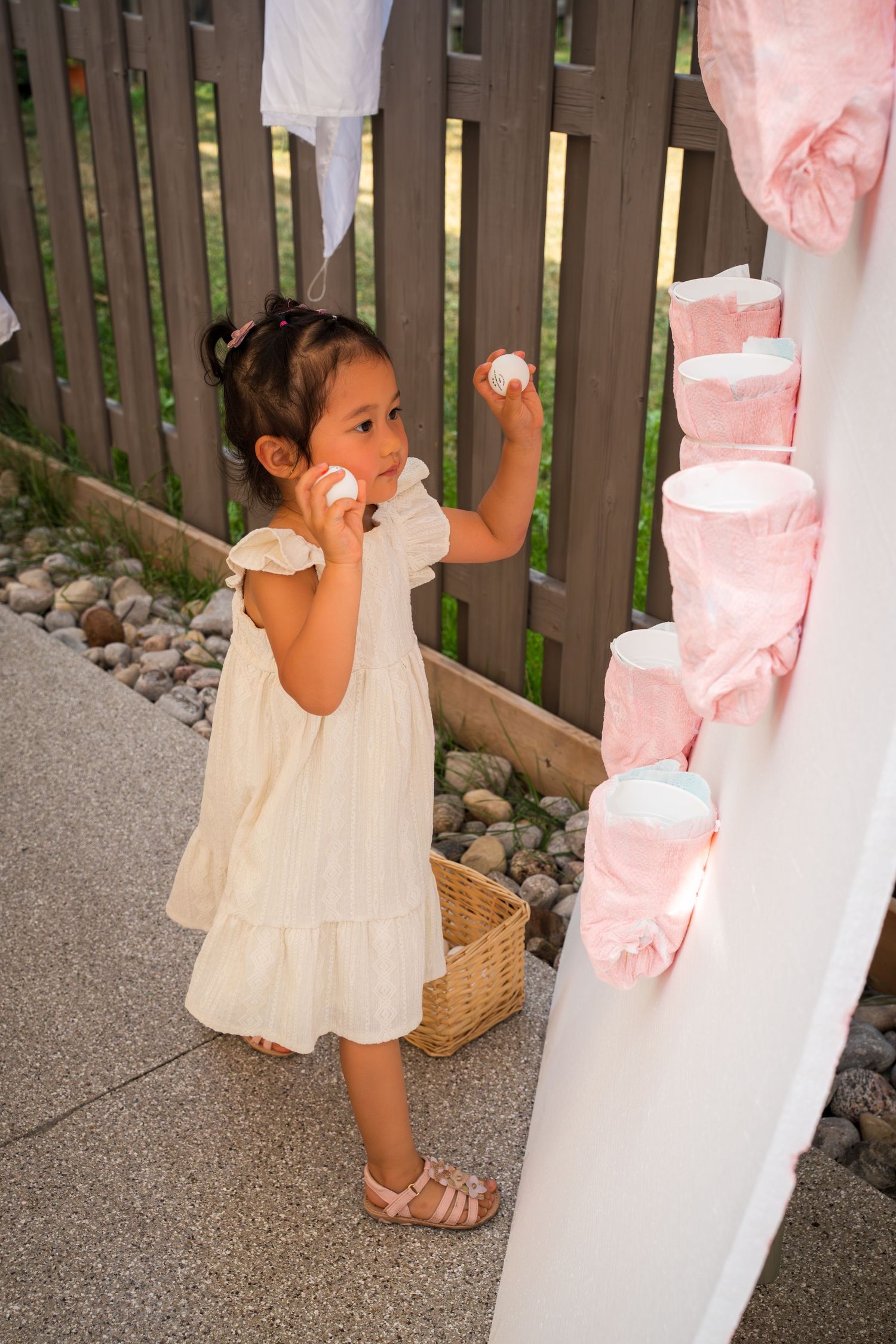 Small child in cream dress playing with pink decorations at an outdoor event.