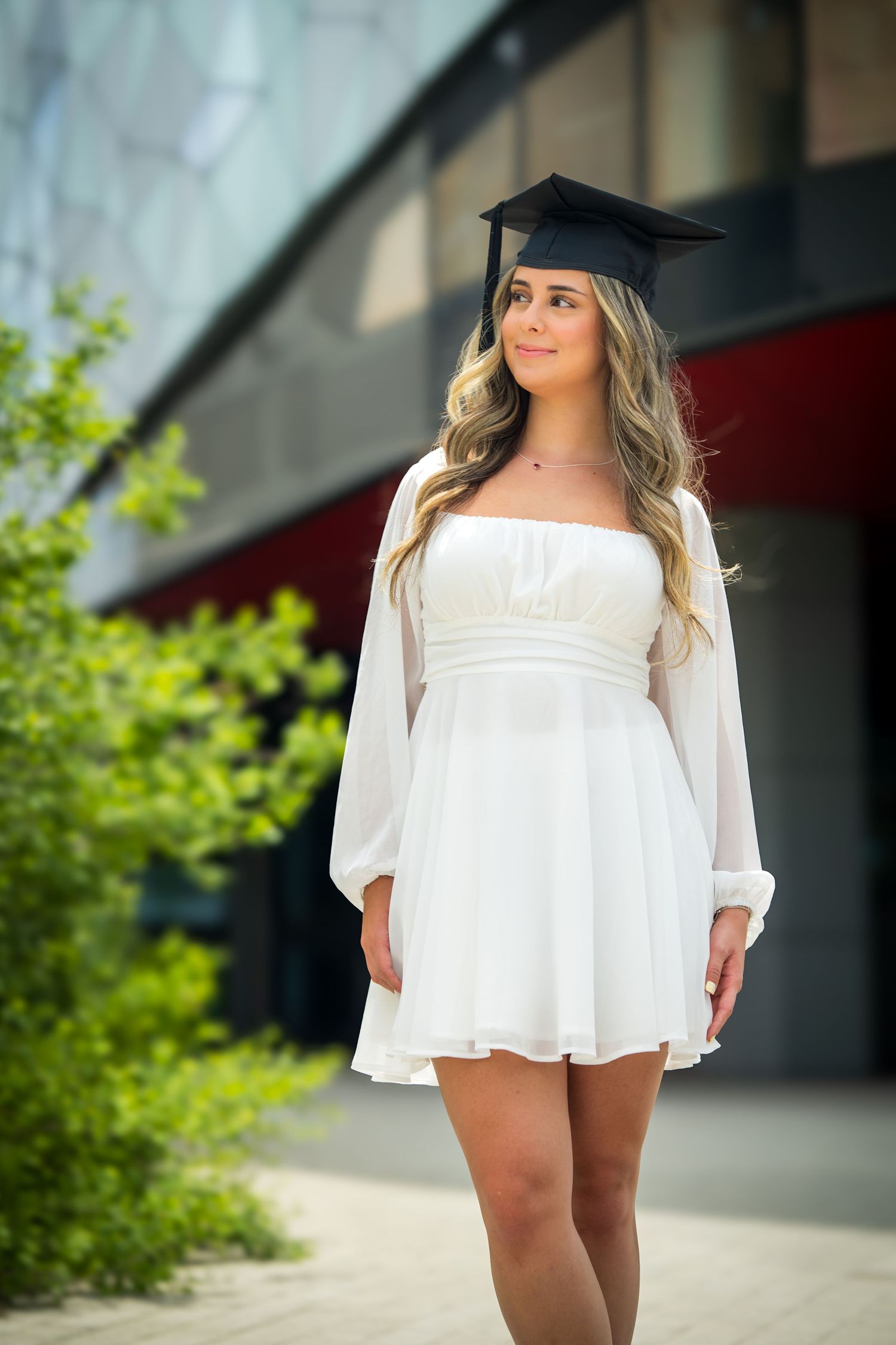 Graduate in flowing white dress poses near modern building and greenery.