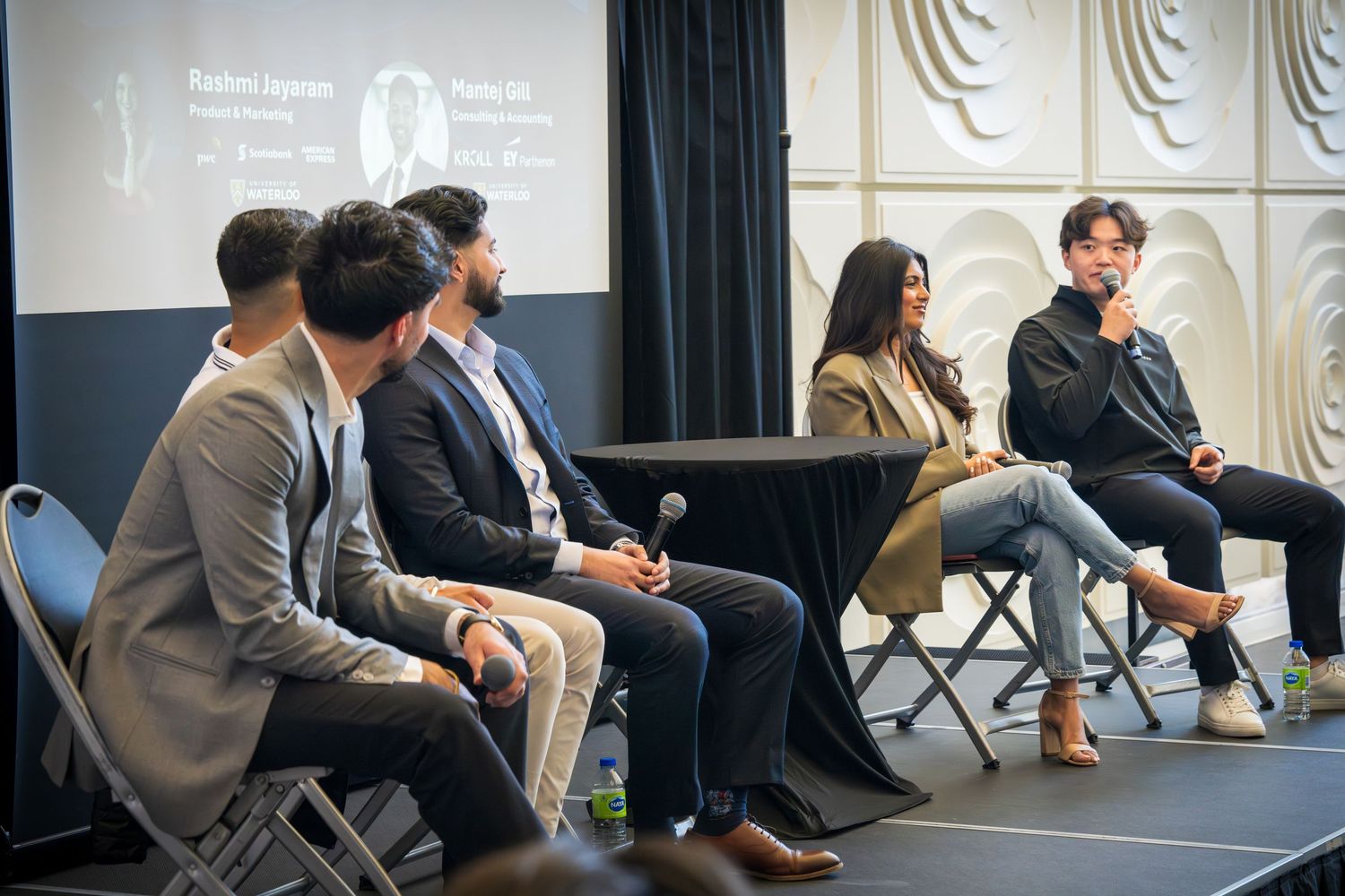 Wide shot of professional panel discussion with speakers seated on stage against textured wall.