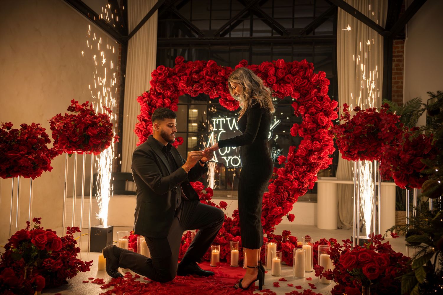 Romantic proposal setup with red rose wall and neon sign illuminated by sparklers and candlelight.