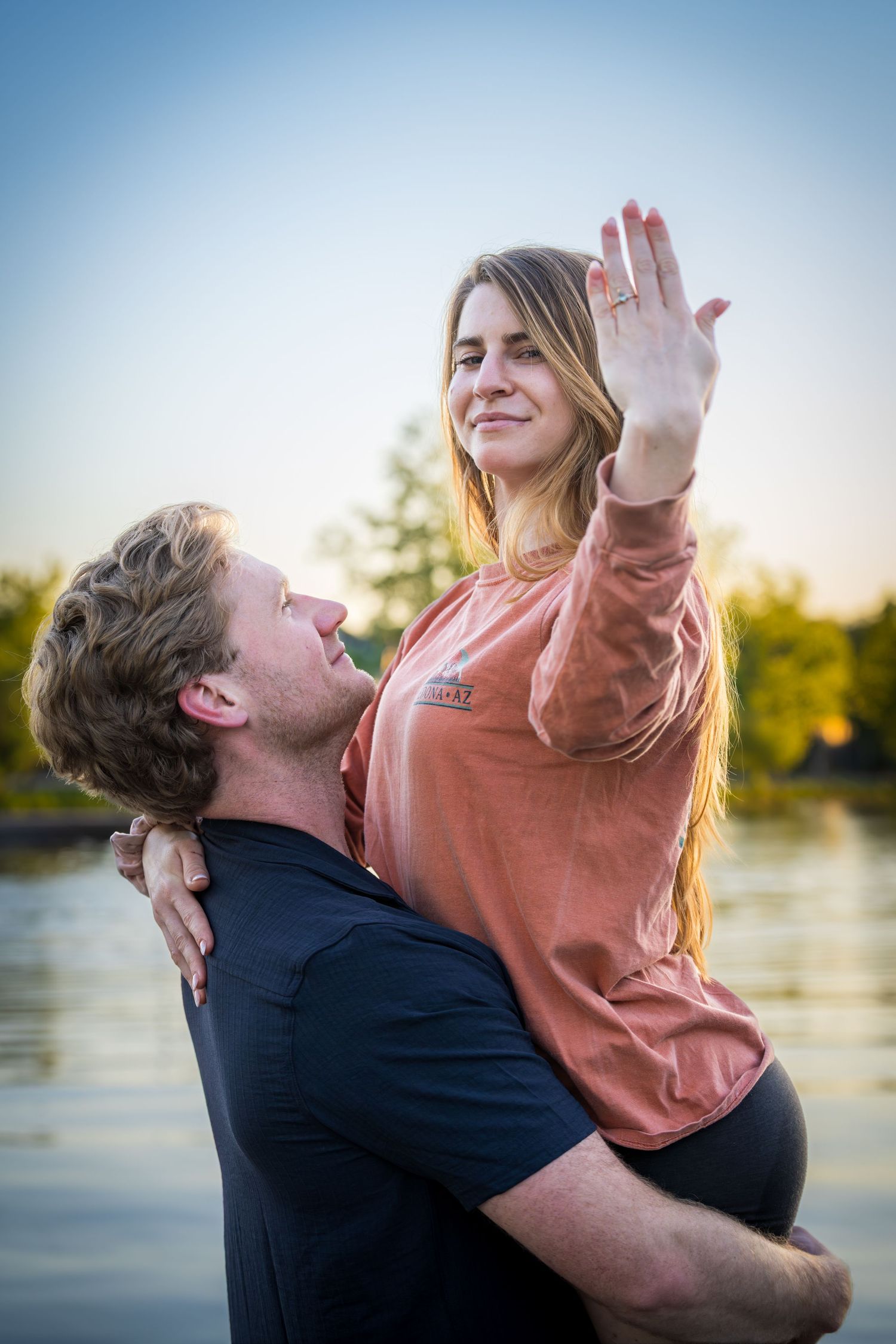 Man lifting woman in coral sweatshirt near a lake at sunset, she shows off her engagement ring with a smile.