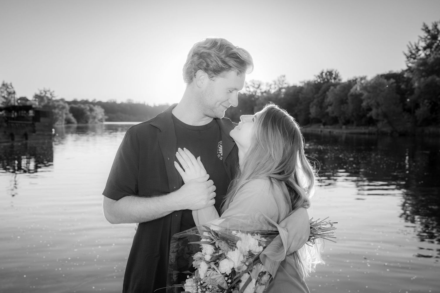 Black and white photo of a couple sharing a romantic moment by a lake at sunset, woman holding flowers.