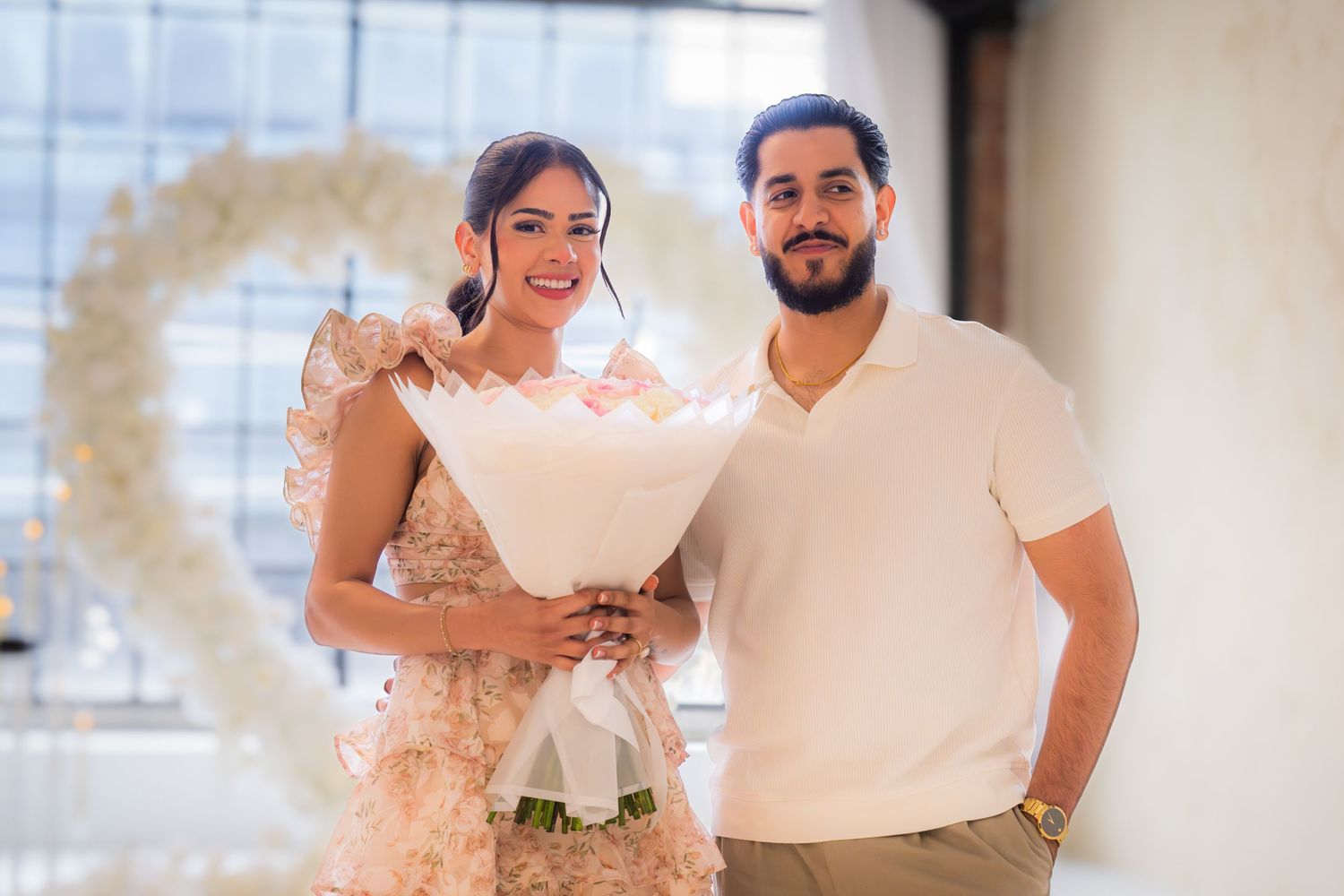 A smiling couple poses together holding a pink and white rose bouquet in front of a floral heart.