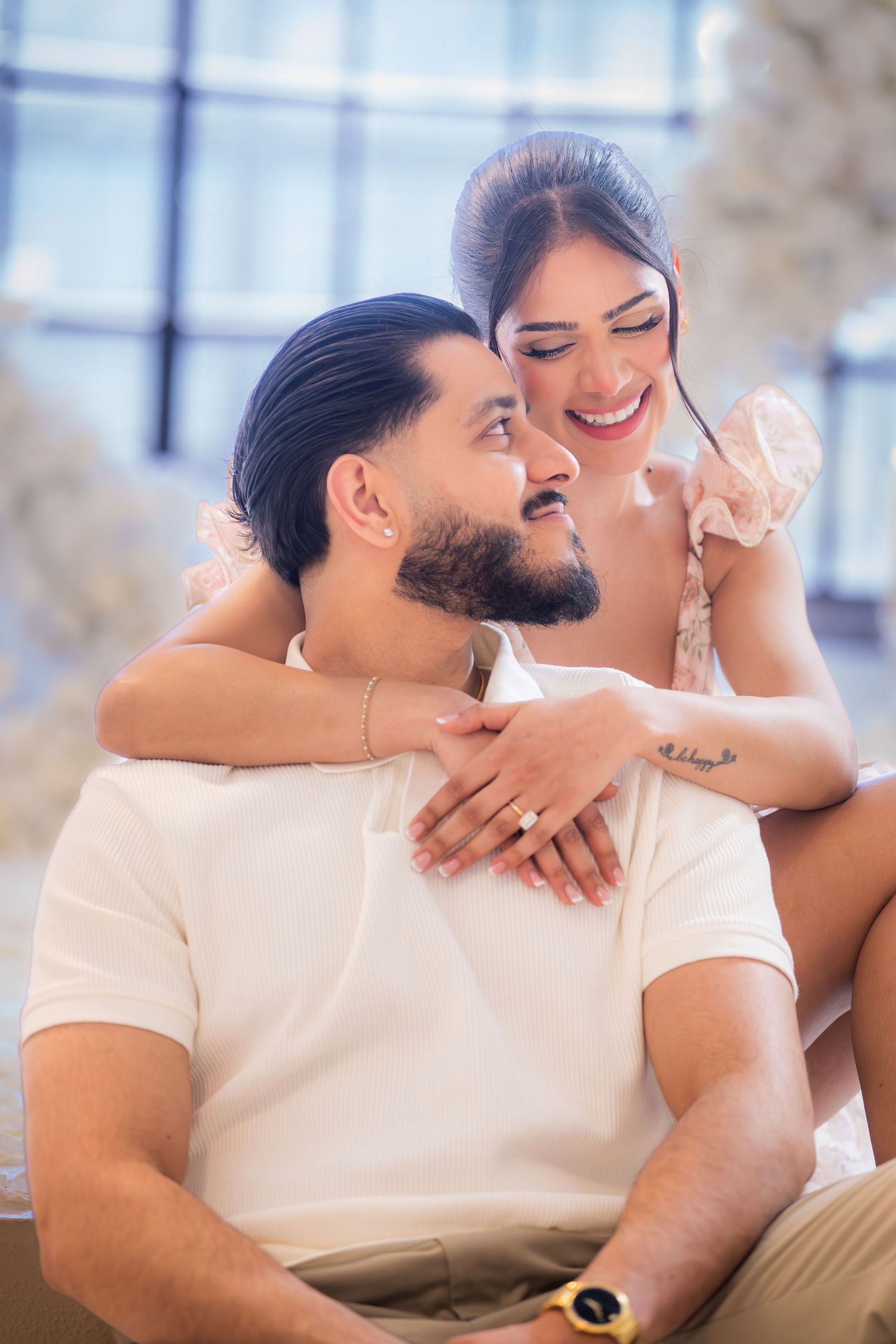 Happy engaged couple embracing, woman showing ring, seated near white floral decor.