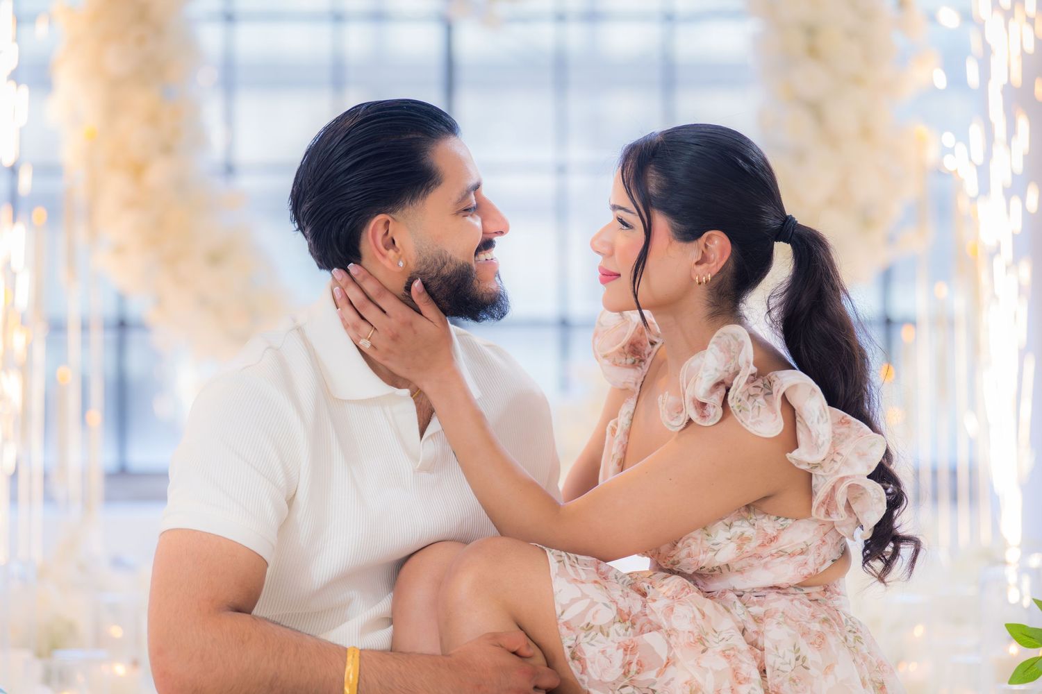 Couple sharing a tender moment surrounded by elegant white floral decorations and lights.
