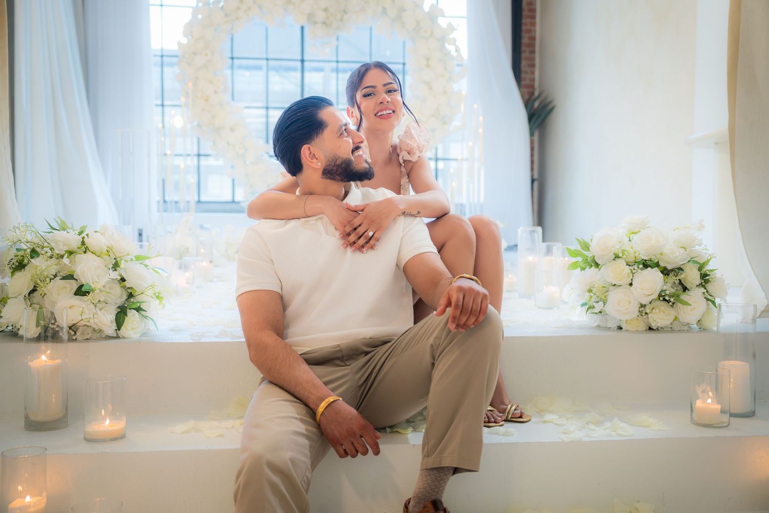 Engaged couple sitting together in front of white floral arch surrounded by candles and flowers.