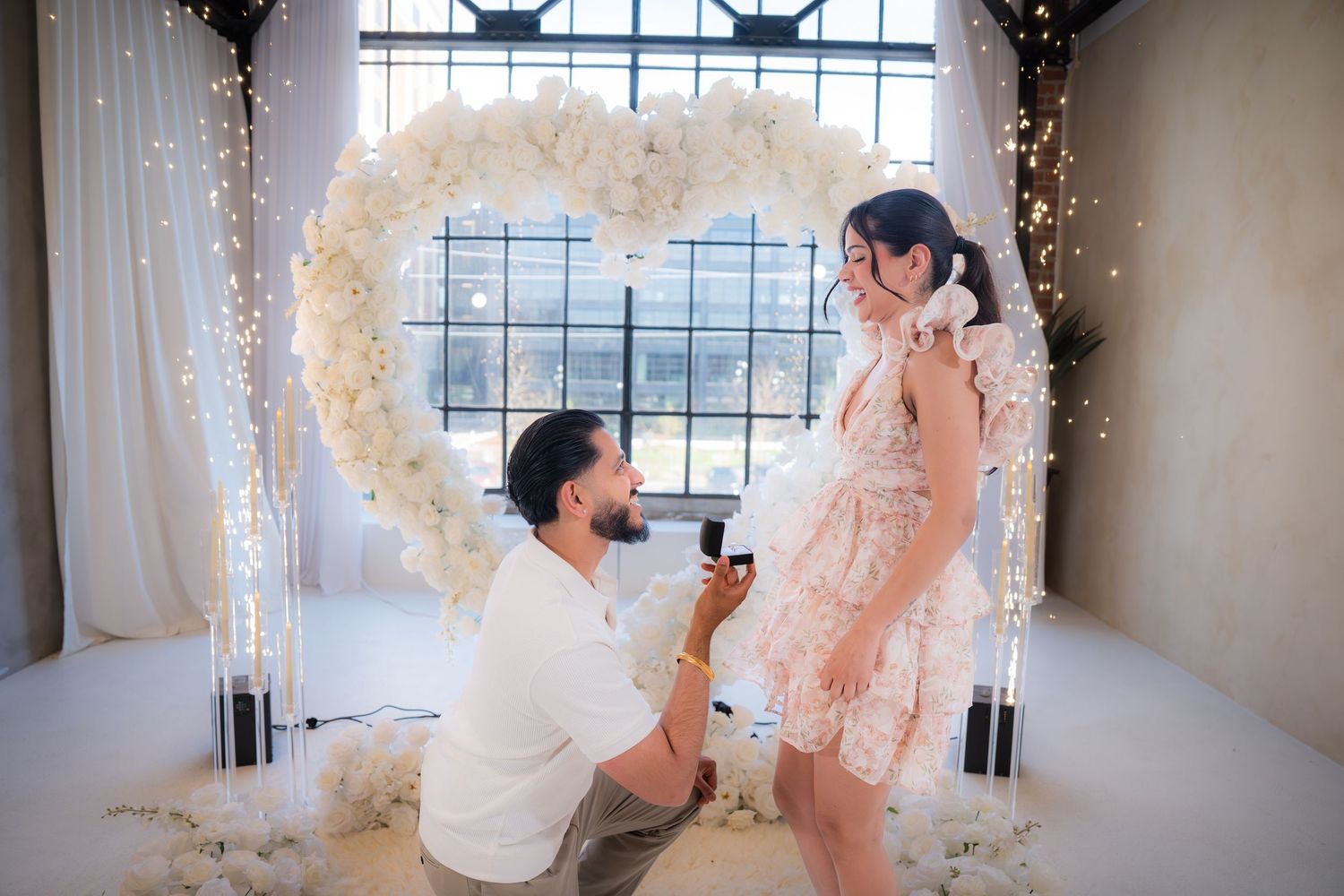 A man proposes to a woman in a floral dress beneath a heart-shaped floral arch with fairy lights.