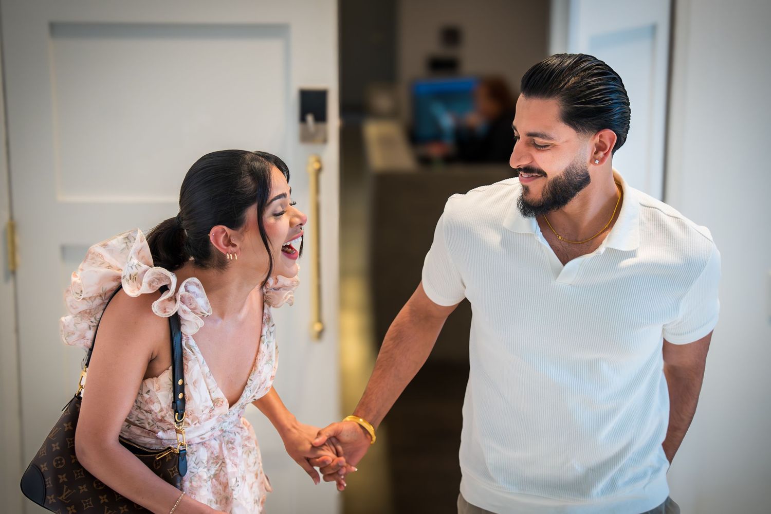 Happy couple holding hands and laughing together, woman in ruffled floral dress, man in white shirt.