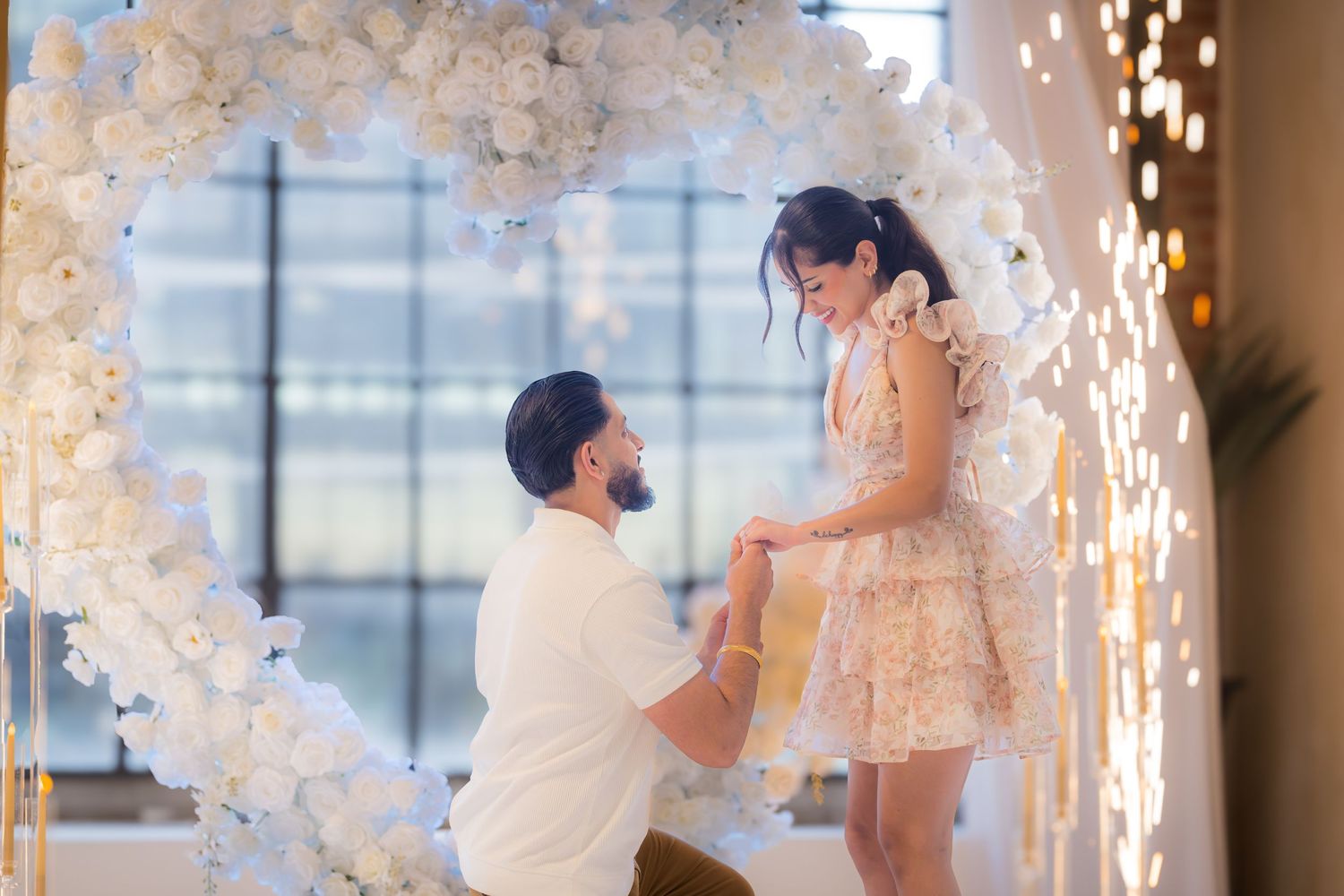 Man proposing on one knee to woman under stunning white floral heart arch at engagement.